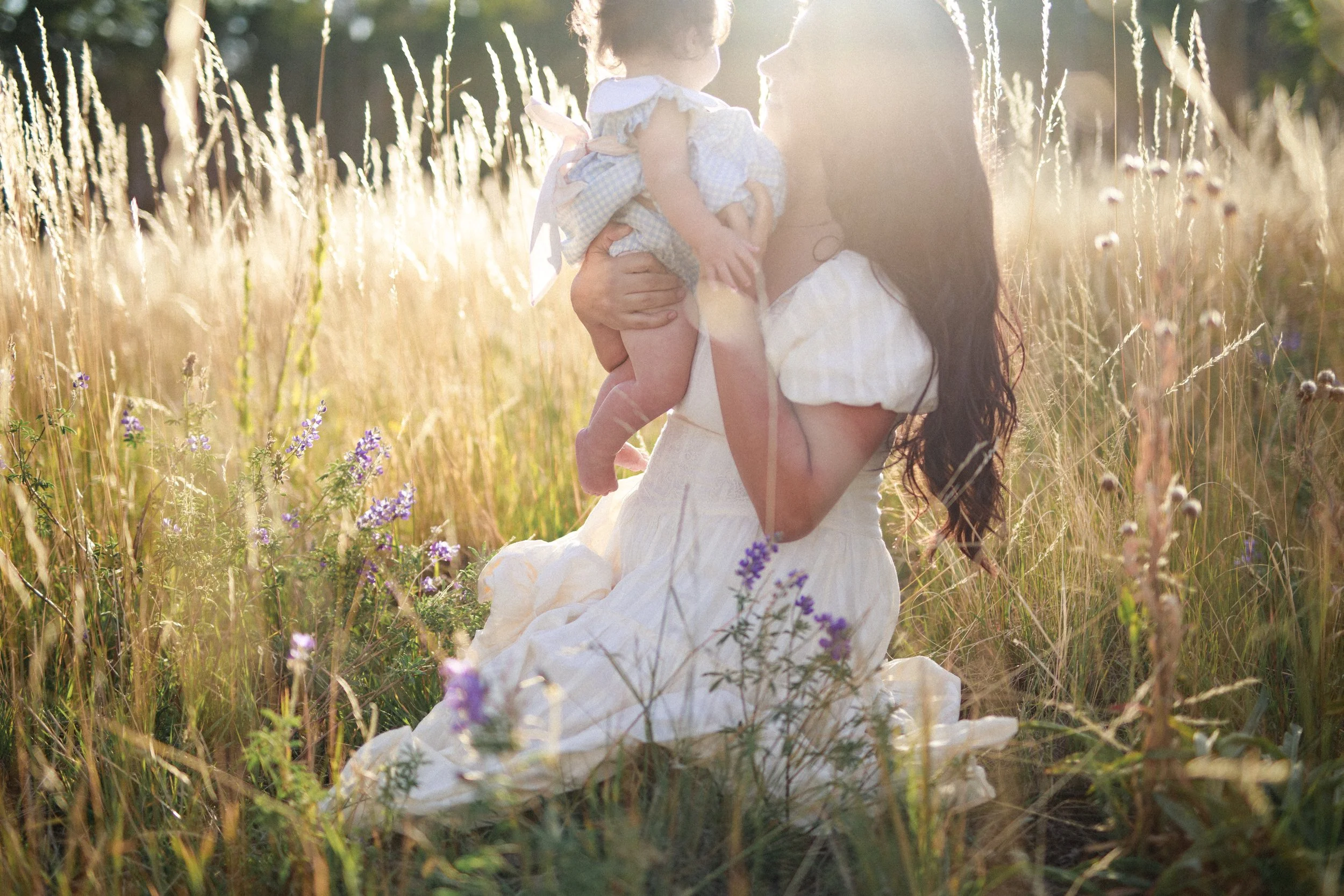 A woman in a white dress kneeling in a field holding a young girl, both backlit by sunlight, surrounded by tall grass and purple wildflowers.