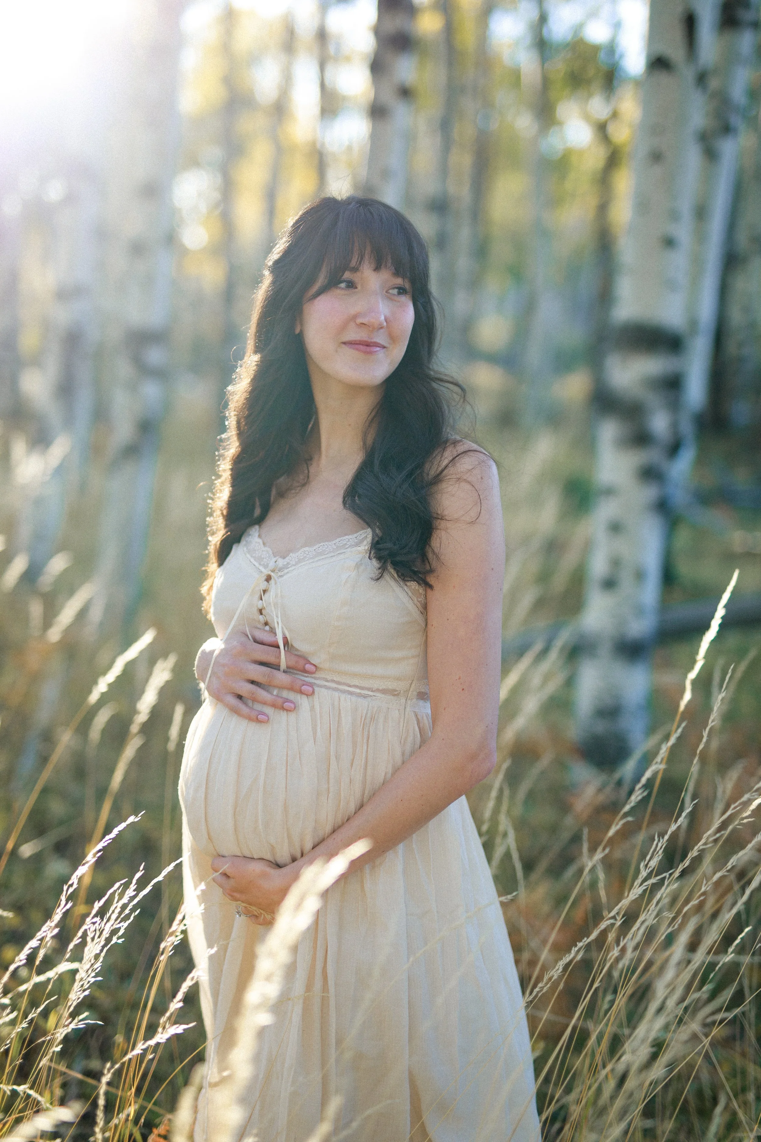 Pregnant woman in a beige dress standing in a forest with trees and tall grasses, holding her belly with one hand and touching her belly with the other, bathed in soft sunlight.