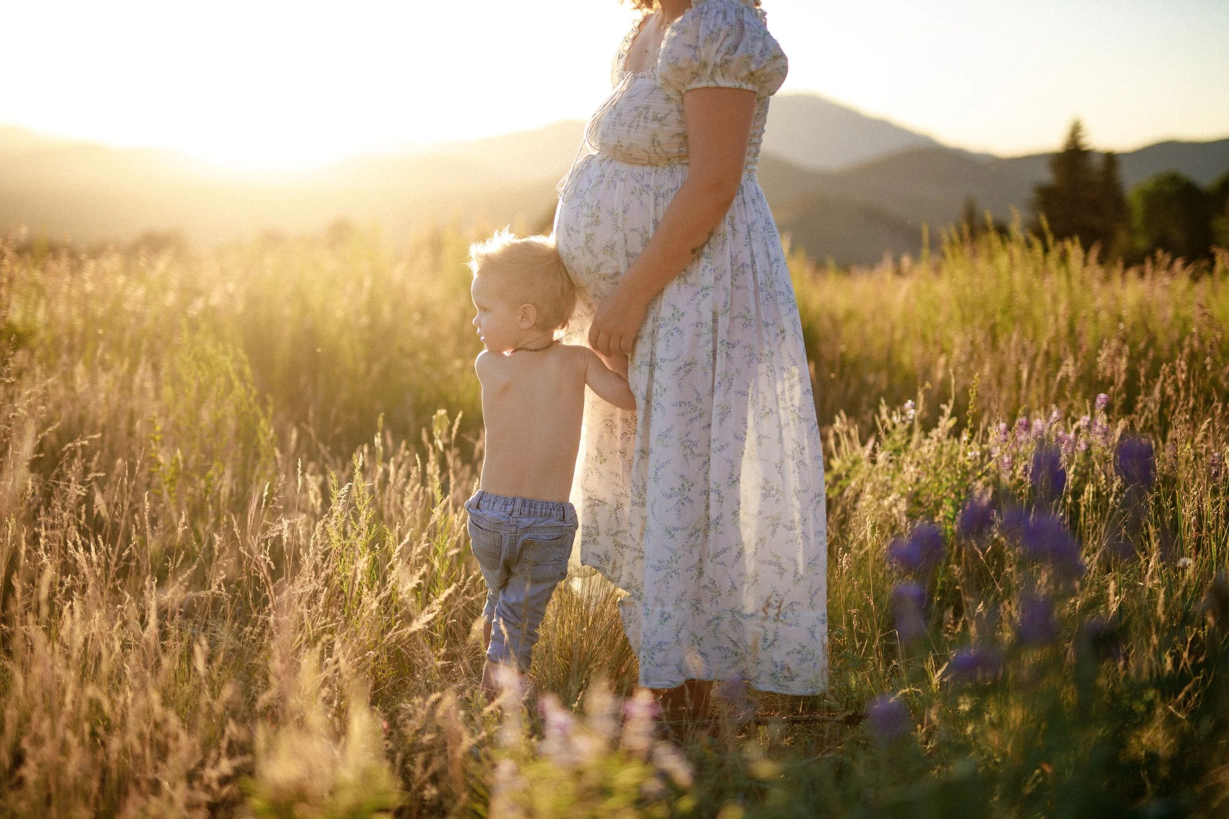 A pregnant woman in a long white floral dress holding the hand of a young shirtless boy in a field of tall grass and wildflowers during sunset.