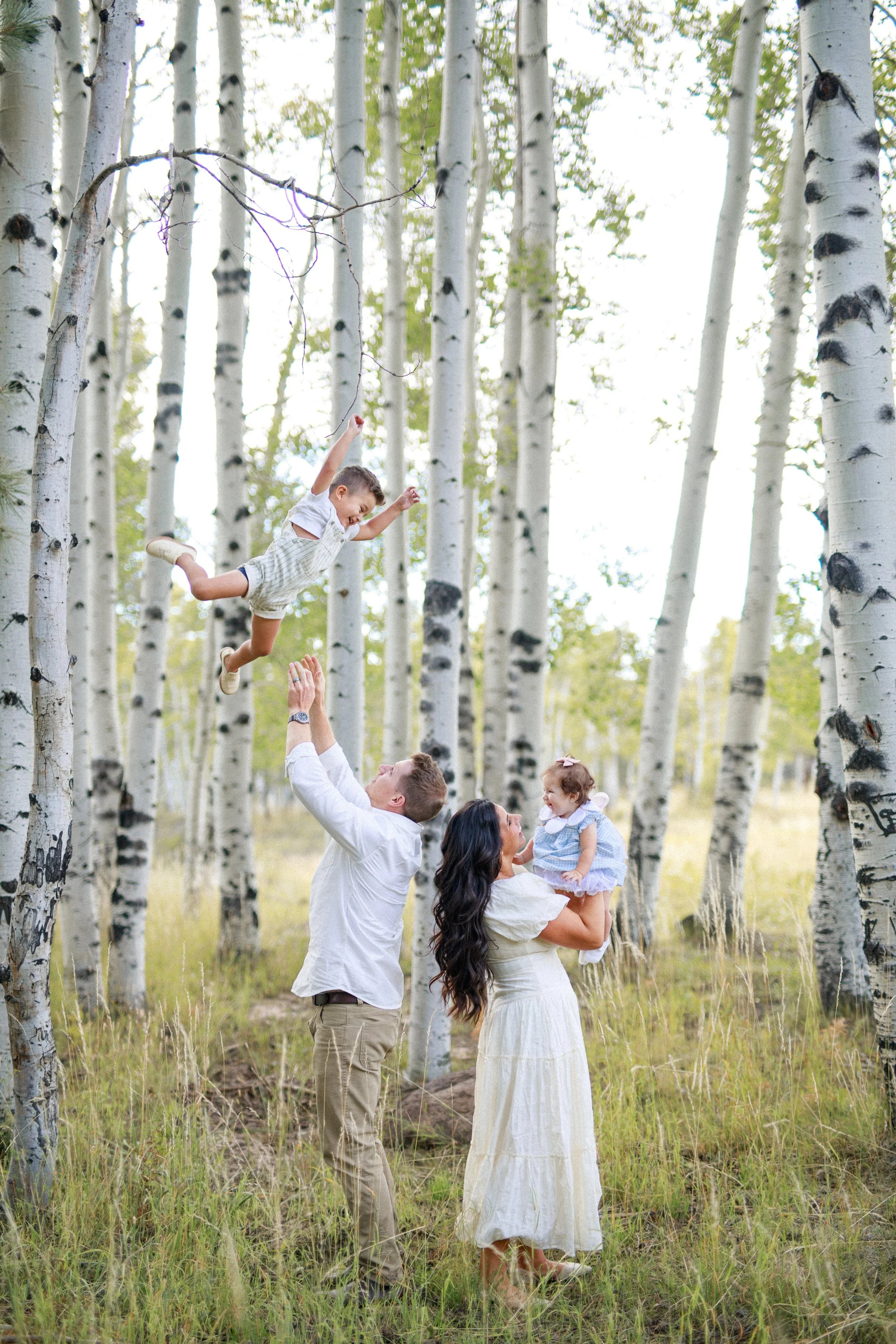 Family of four, consisting of a father, mother, and two children, enjoying time in a forest of tall, white-barked aspen trees. The father is tossing a boy into the air while the mother holds a girl, and the boy is mid-air with outstretched arms and a