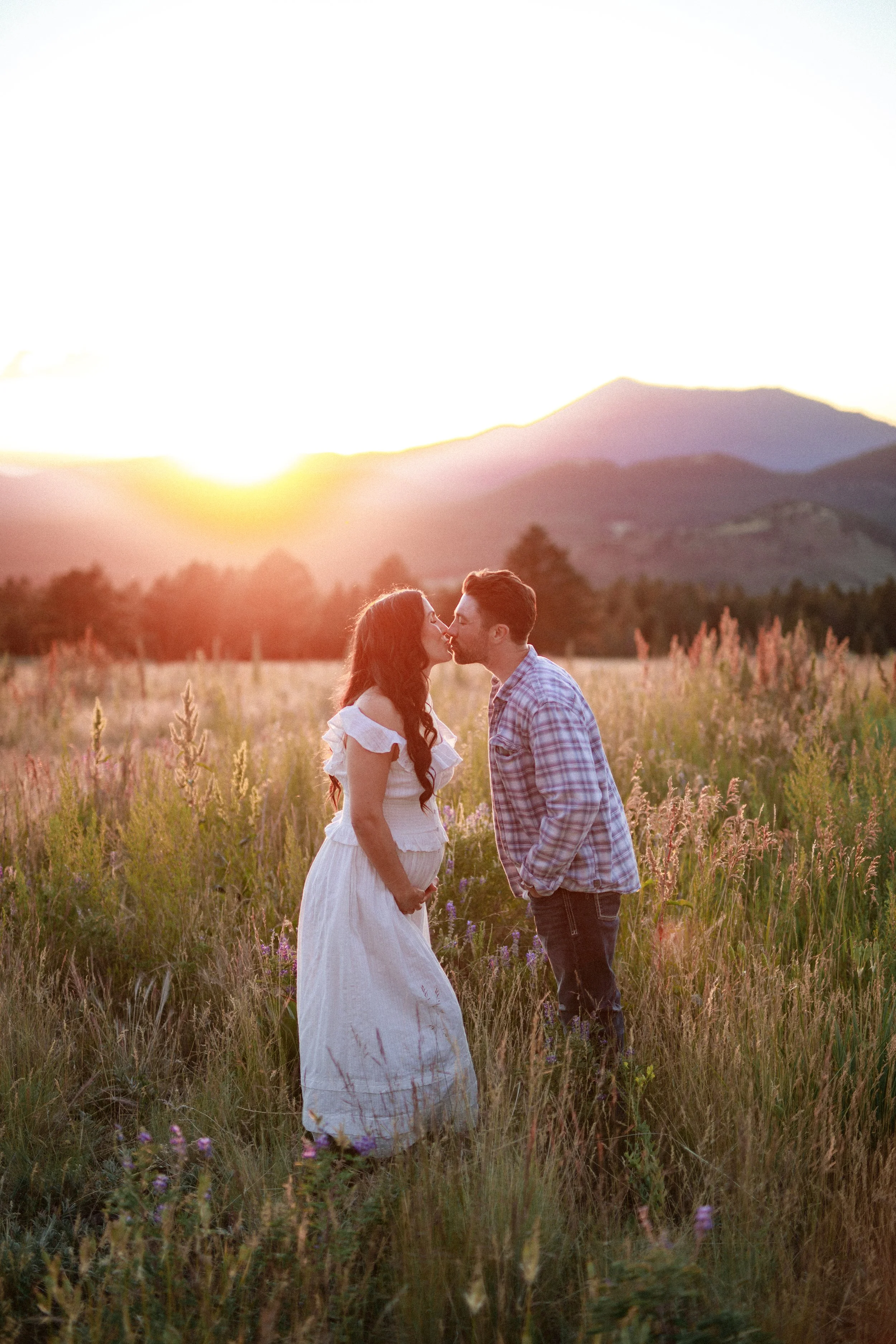 Couple standing in a field of wildflowers at sunset, about to kiss, with mountains in the background.