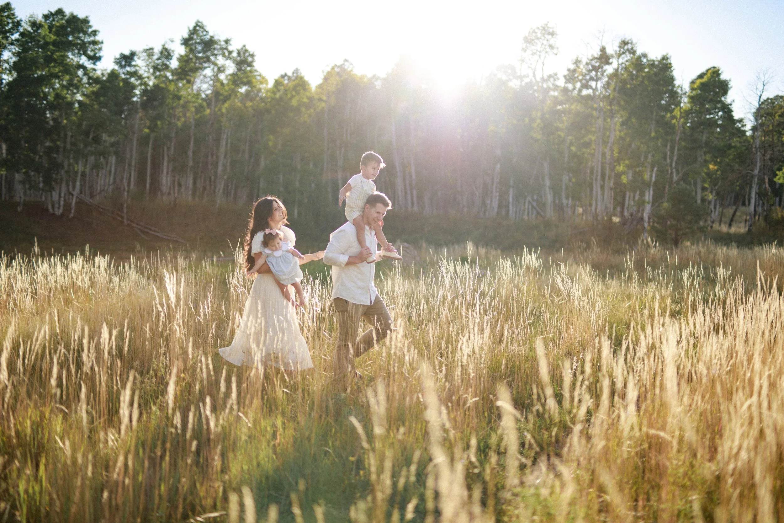 A family of four walking through a grassy field during sunset. The father carries a child on his shoulders, the mother holds a baby, and the child on his shoulders looks ahead. The scene is lit by warm sunlight with trees in the background.