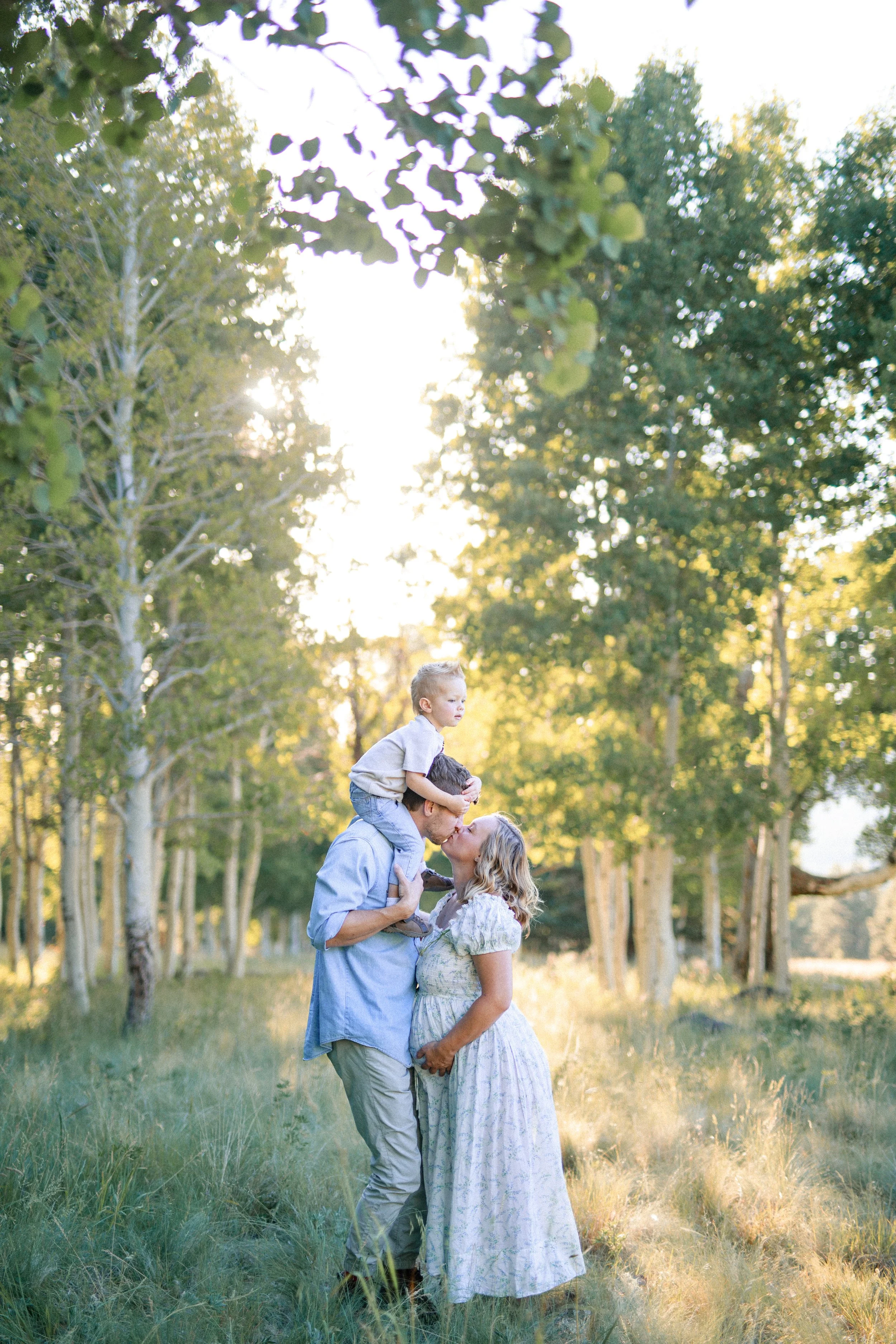 A family of three sharing a kiss outdoors in a forest with tall trees and sunlight shining through the leaves.