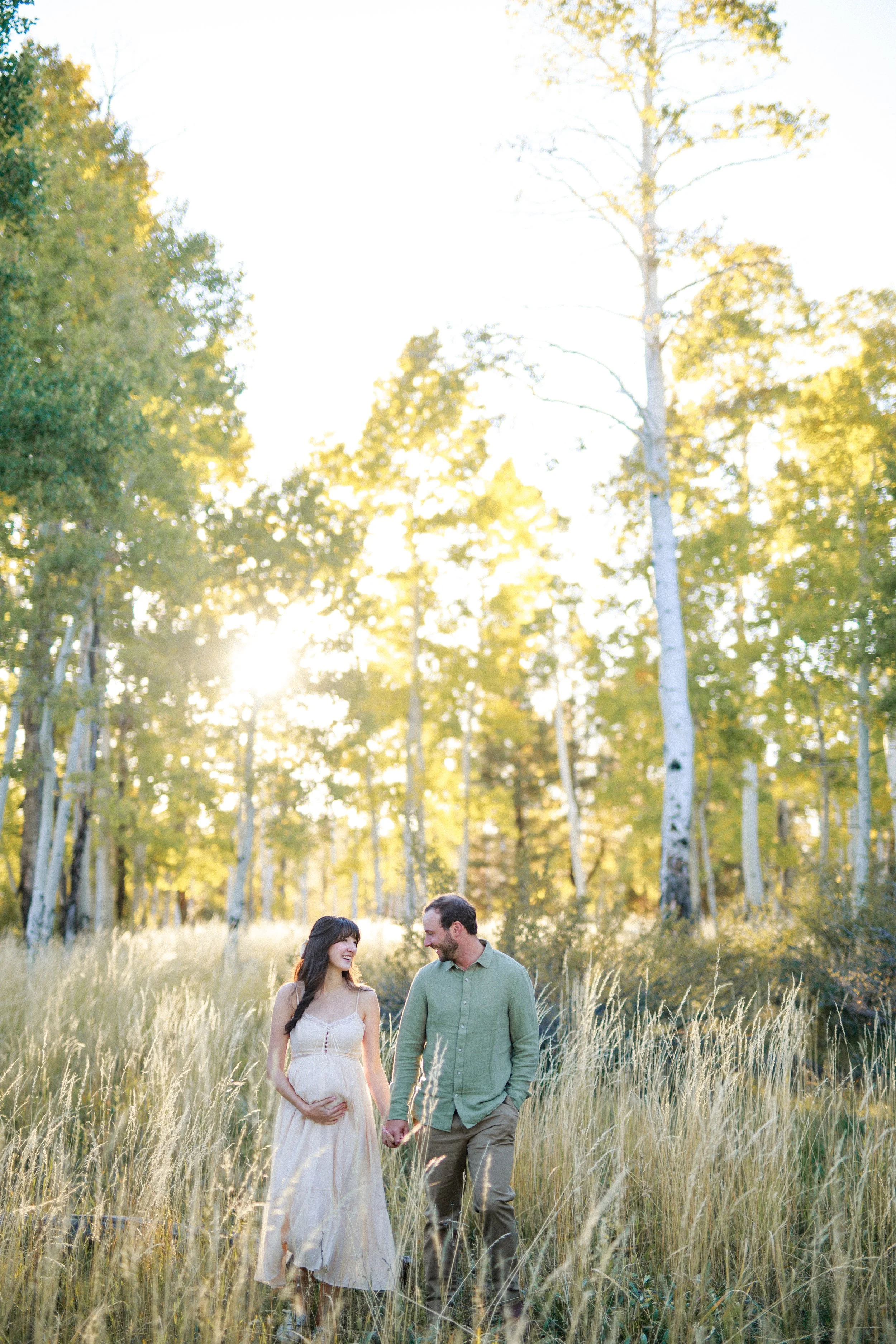 A couple walking hand in hand through a grassy field during sunset, surrounded by trees.