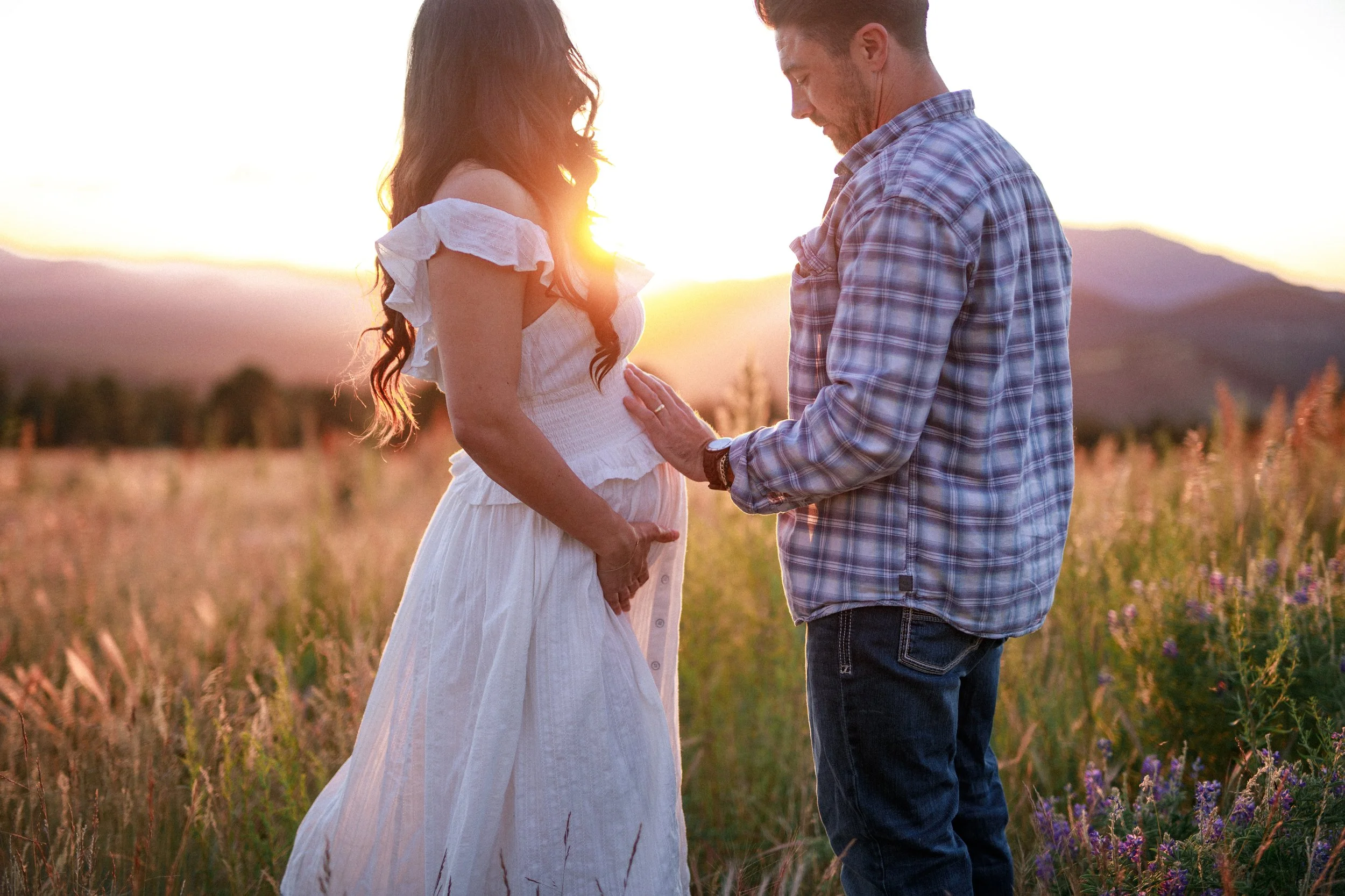 A couple stands in a field during sunset, with the woman pregnant, touching her belly while the man gently places his hand on her abdomen.