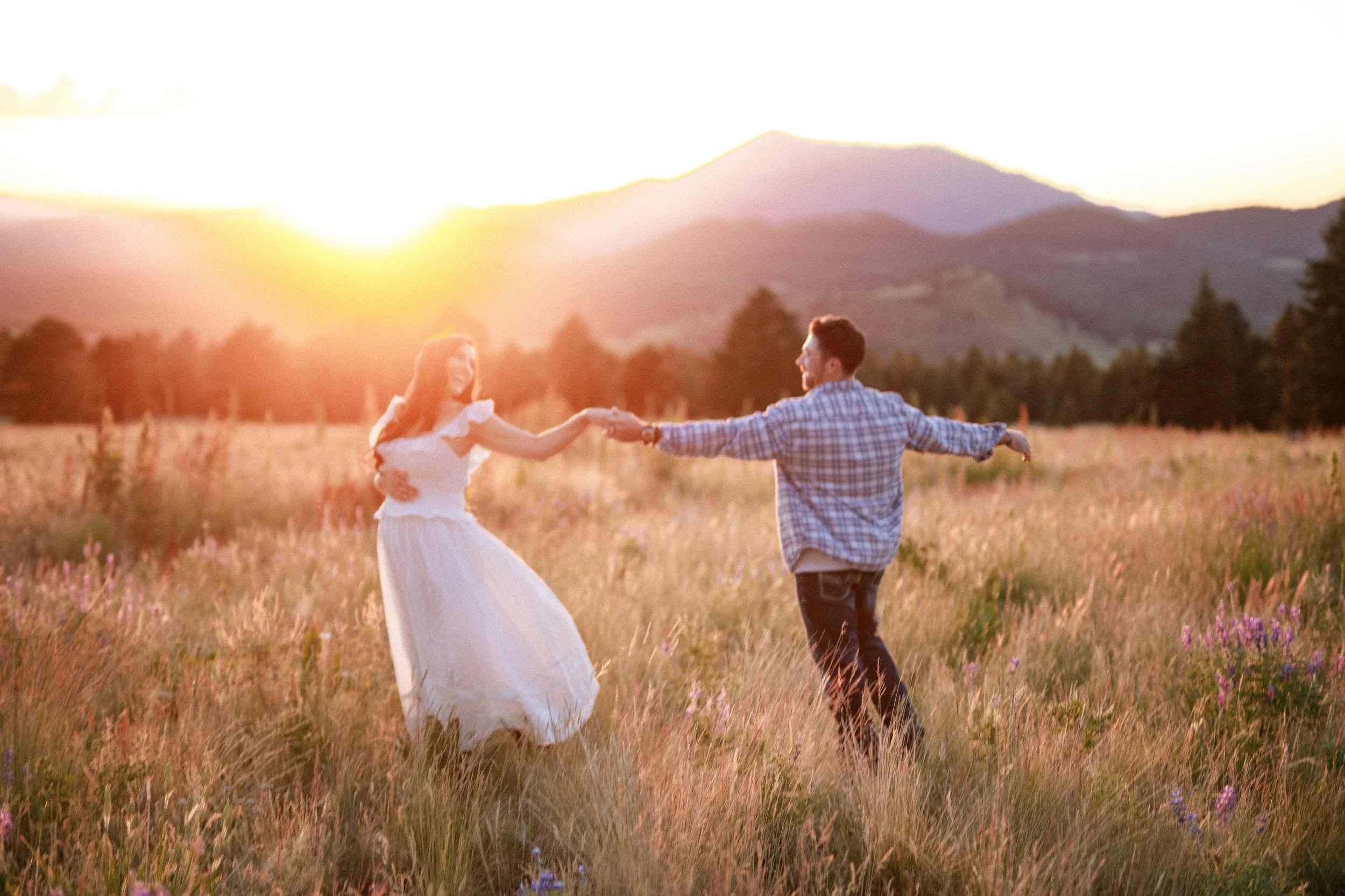 A couple dancing in a grassy field at sunset with mountains in the background.