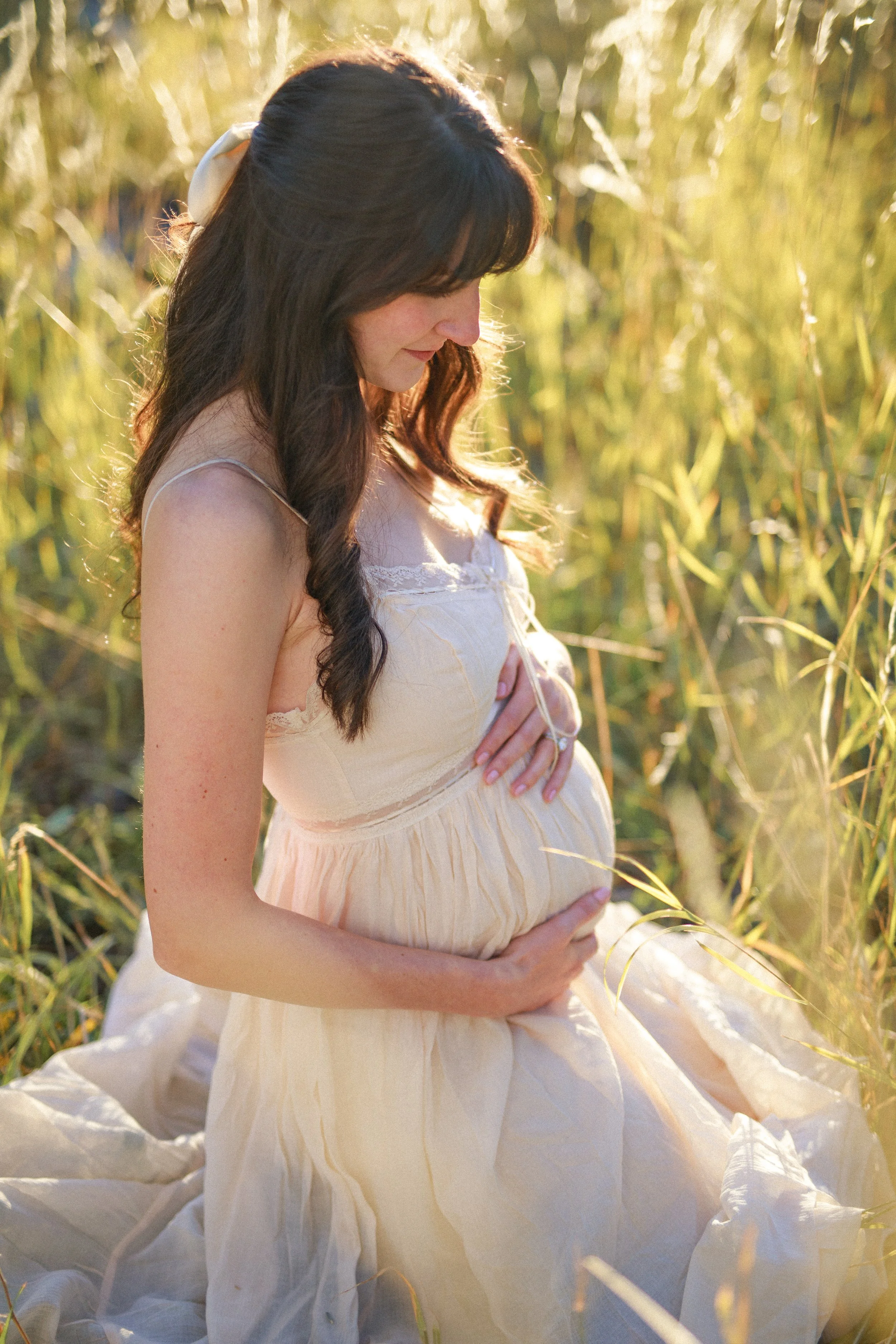 A pregnant woman with dark brown hair, wearing a light cream dress, sitting in a field of tall grass during golden hour, gently holding her belly and smiling softly.