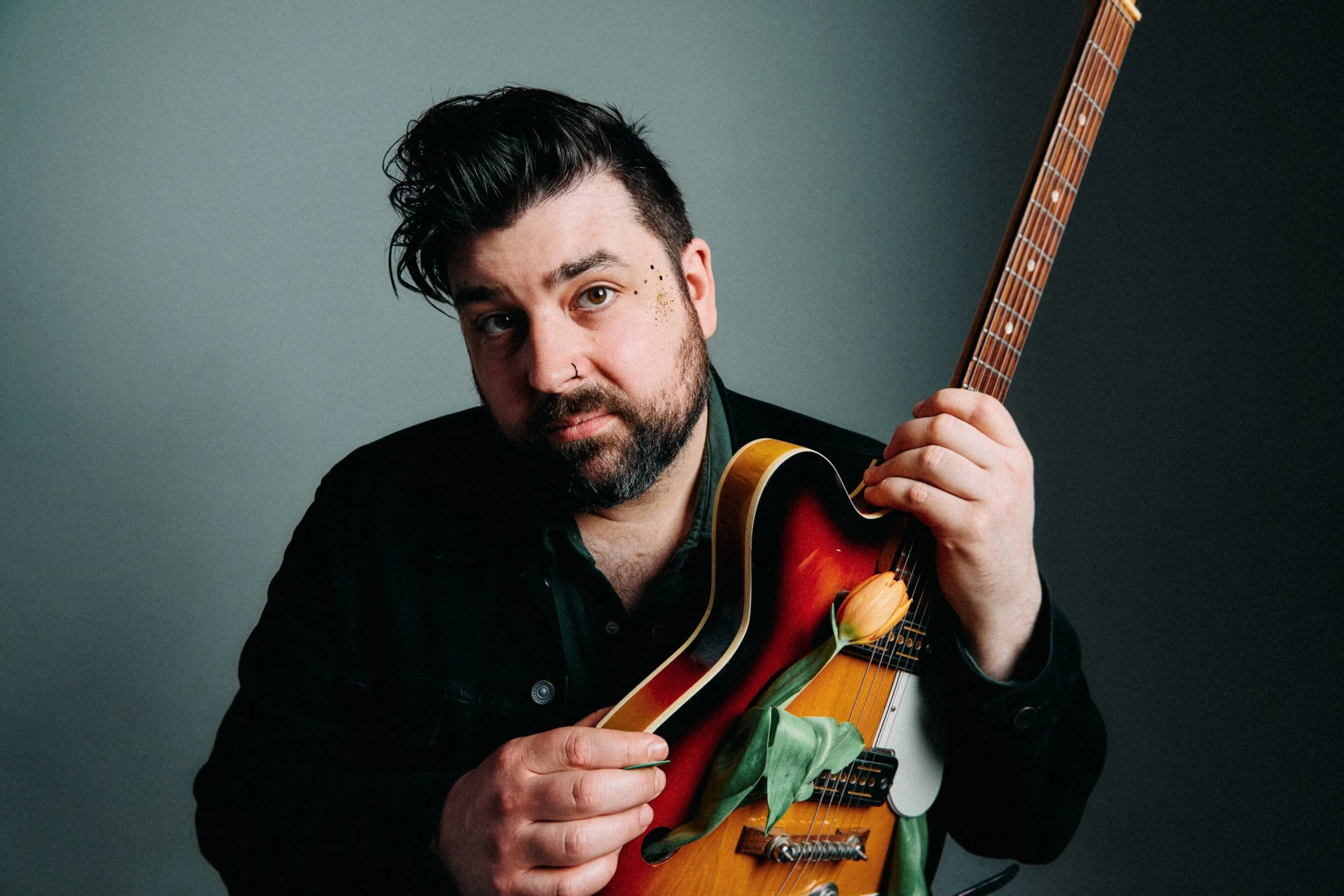 Studio portrait of singer-songwriter Erik Bleich holding a semi-hollow electric guitar with a tulip in the f-hole, photographed against a grey background.
