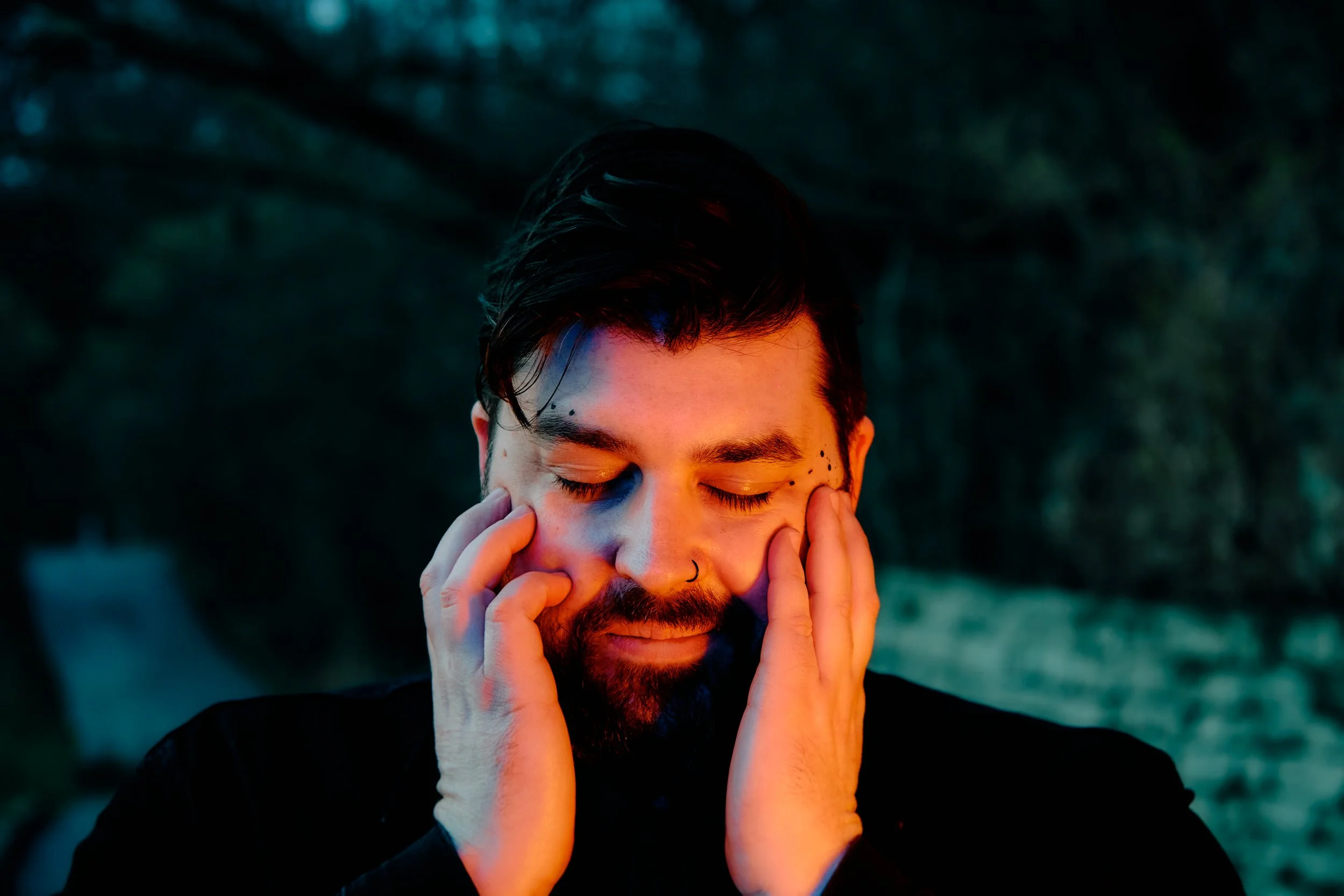 Close-up landscape photo of singer-songwriter Erik Bleich outdoors, smiling with his eyes closed and hands against his face, illuminated by warm and cool light reminiscent of a campfire glow.