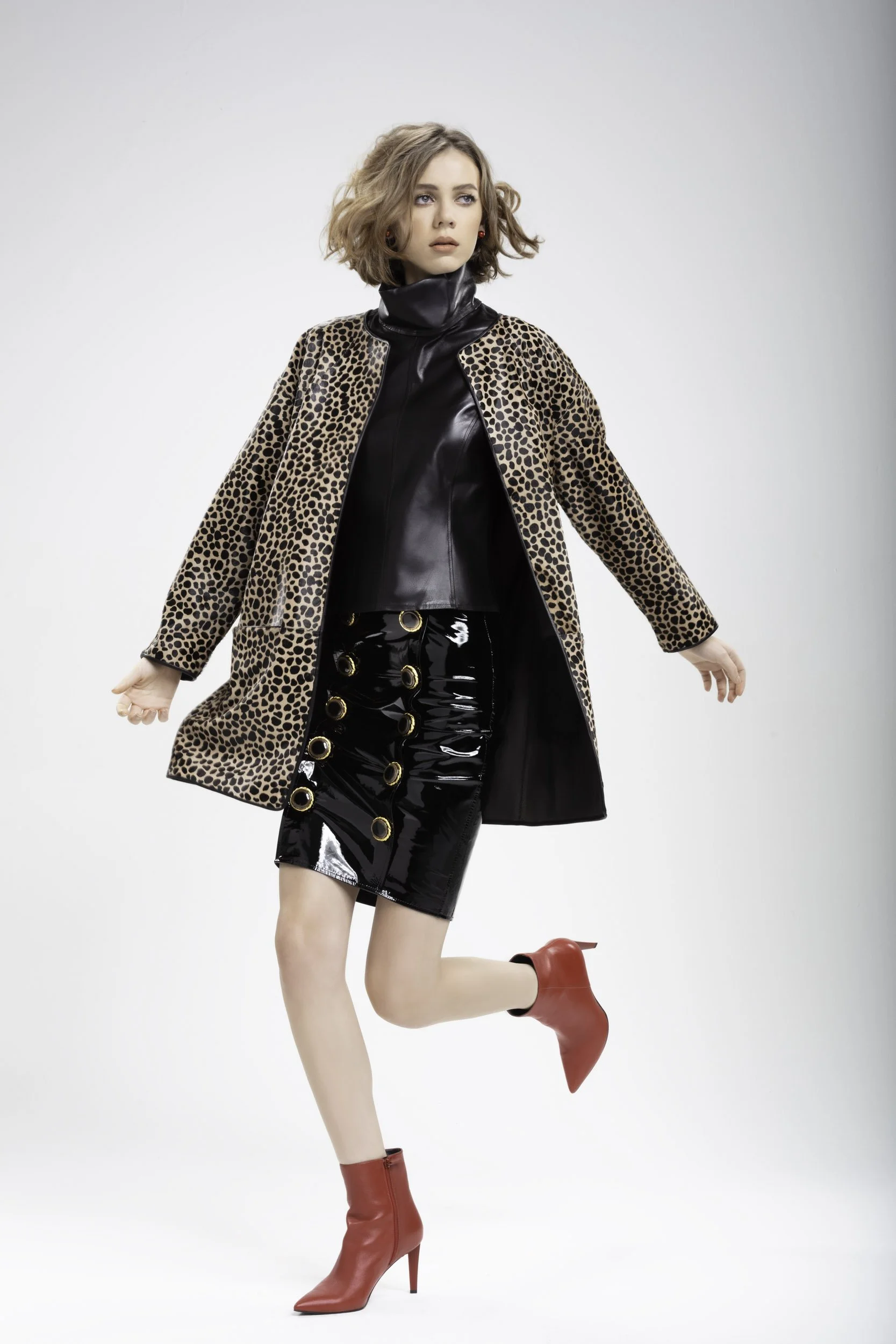 Mujer con cabello rizado, vistiendo abrigo de leopardo, chaqueta de cuero negra, falda de charol negro con anillos dorados, botas rojas de tacón alto en postura de una pierna levantada.