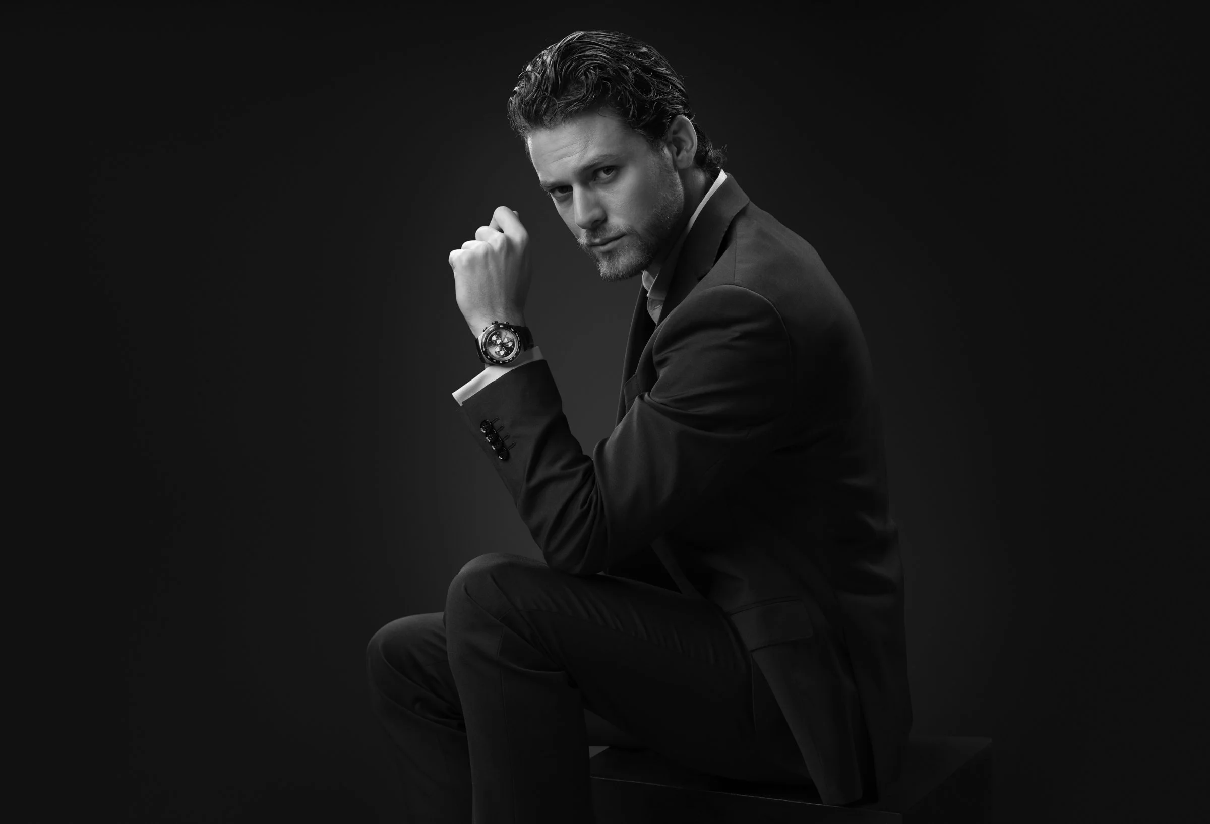 Retrato en blanco y negro de un hombre con cabello rizado peinado hacia atrás, con barba y usando traje elegante, sentado con postura pensativa y mostrando una expresión seria, con un reloj en la muñeca izquierda, en un fondo oscuro.