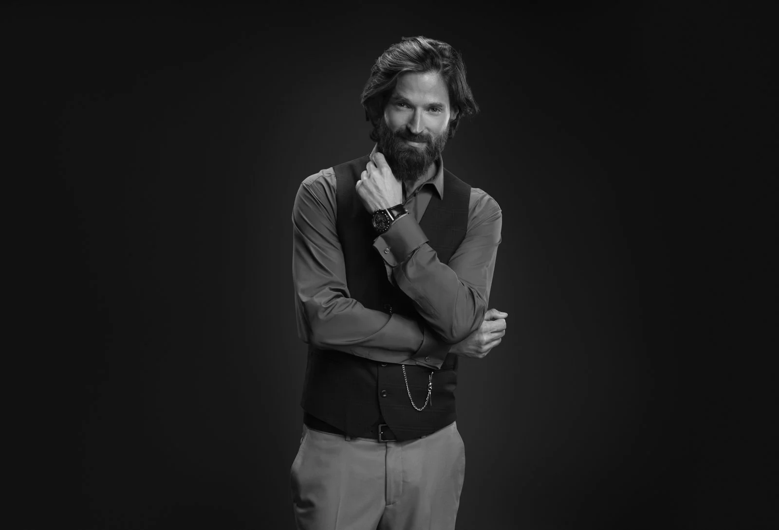 Hombre con barba, cabello largo y vestido formal, con chaqueta y pantalones, posando con brazos cruzados, en un fondo oscuro.