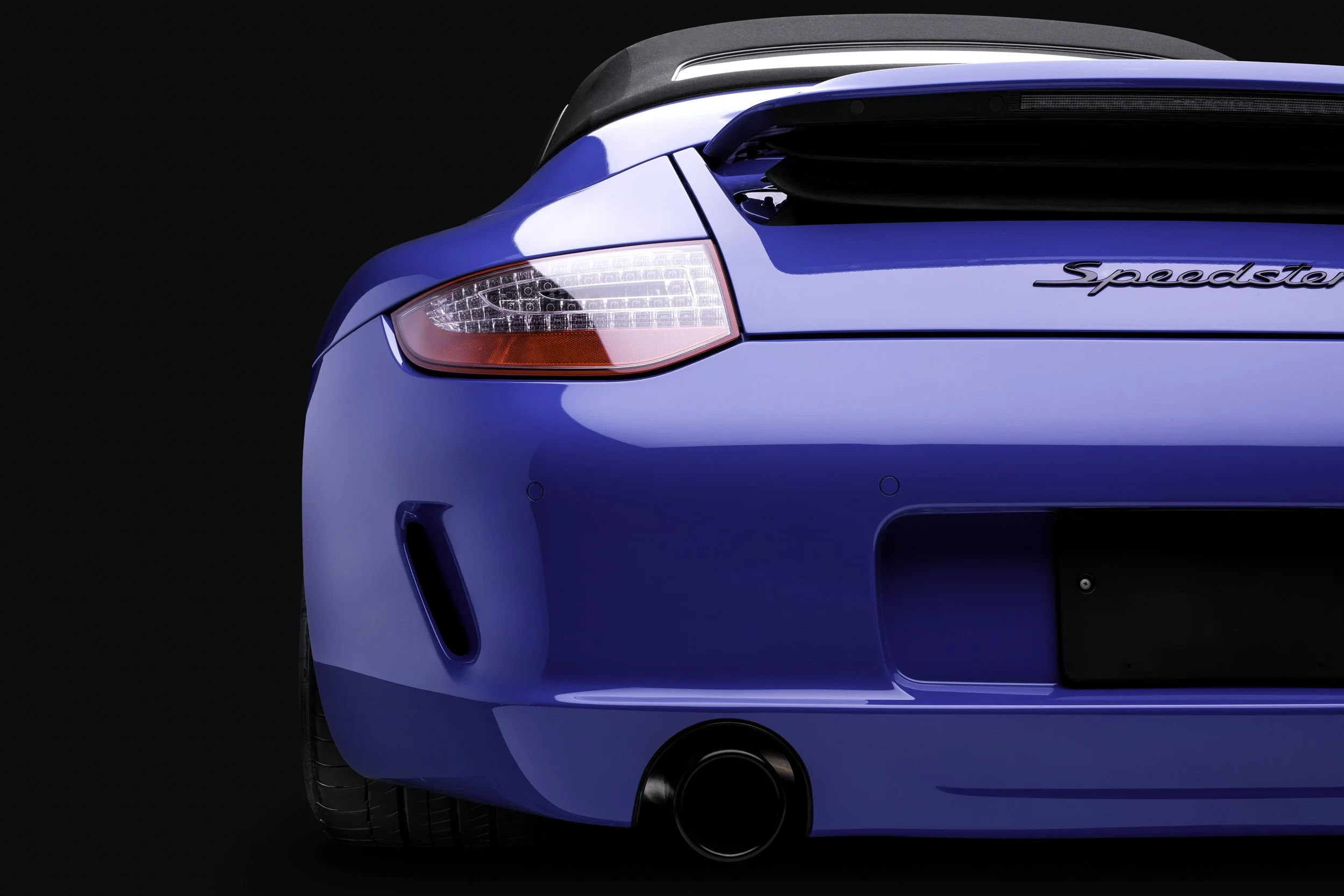 Parte trasera de un automóvil azul con la palabra 'Speedster' en el panel lateral, luz trasera y neumáticos, fondo negro.