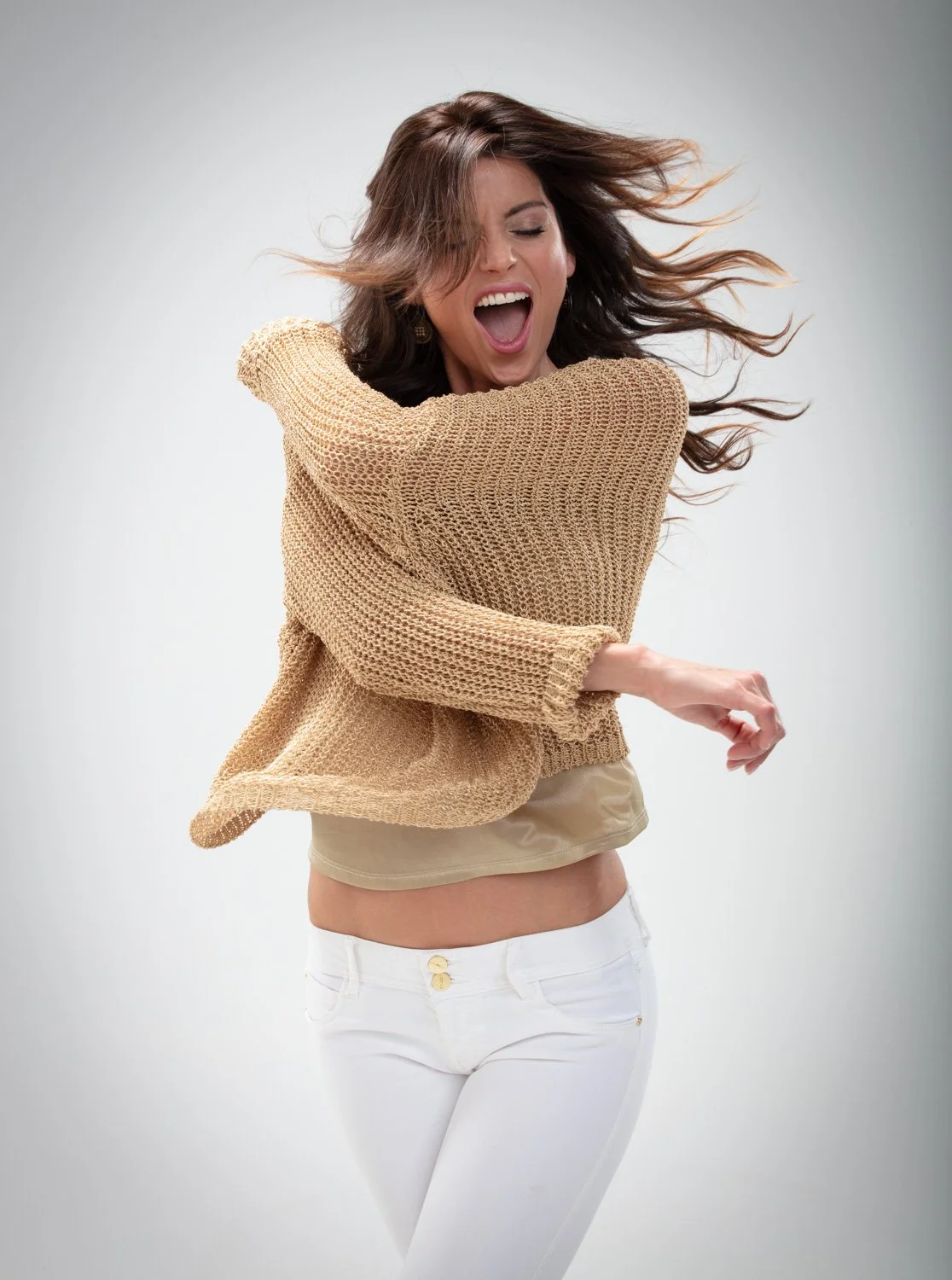 Mujer sonriendo con cabello al viento, vistiendo suéter de hilo y pantalones blancos.
