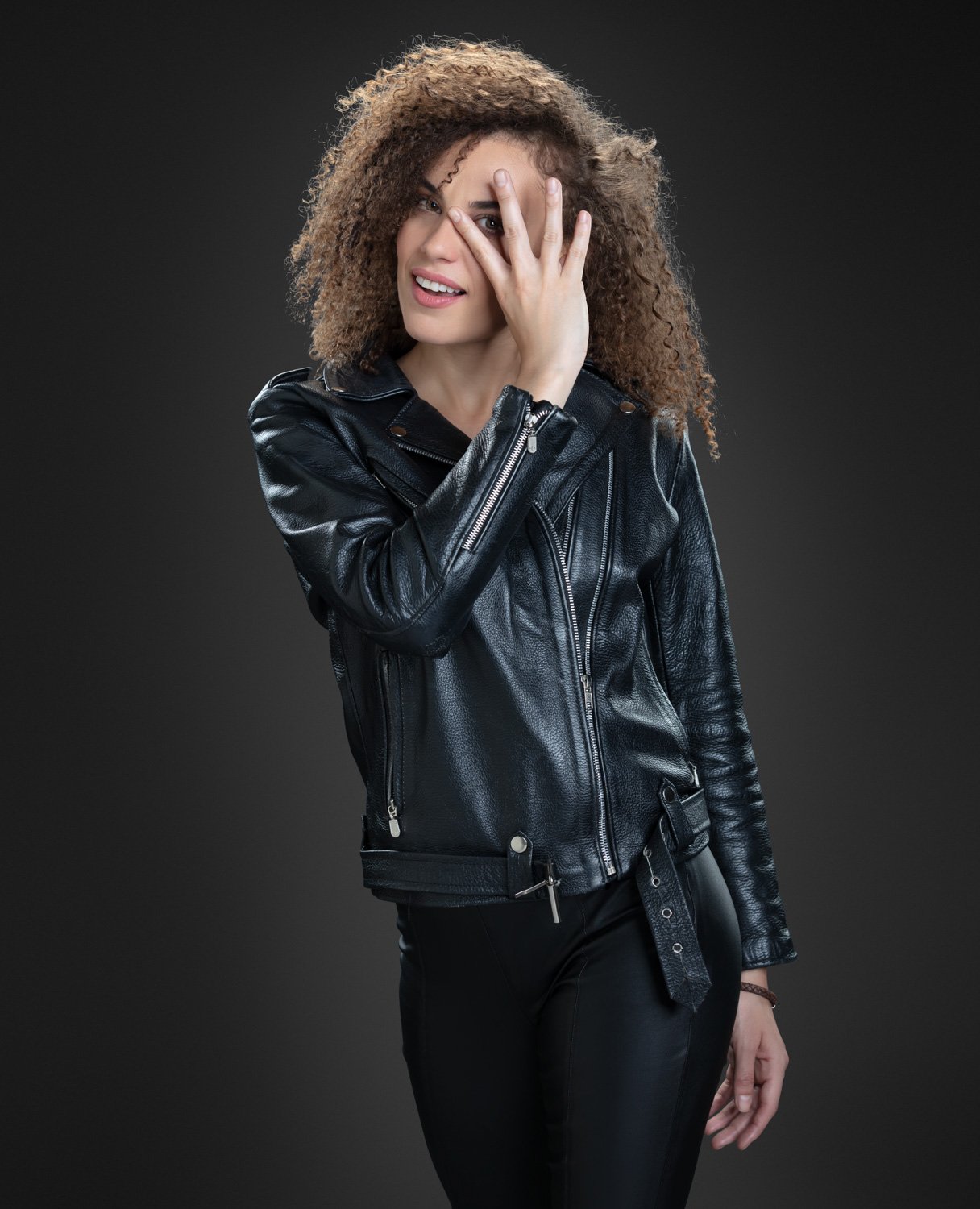 Joven mujer con cabello rizado y rubio, vestido con chaqueta de cuero negra, haciendo un gesto de cubrirse un ojo con la mano en un fondo gris oscuro.