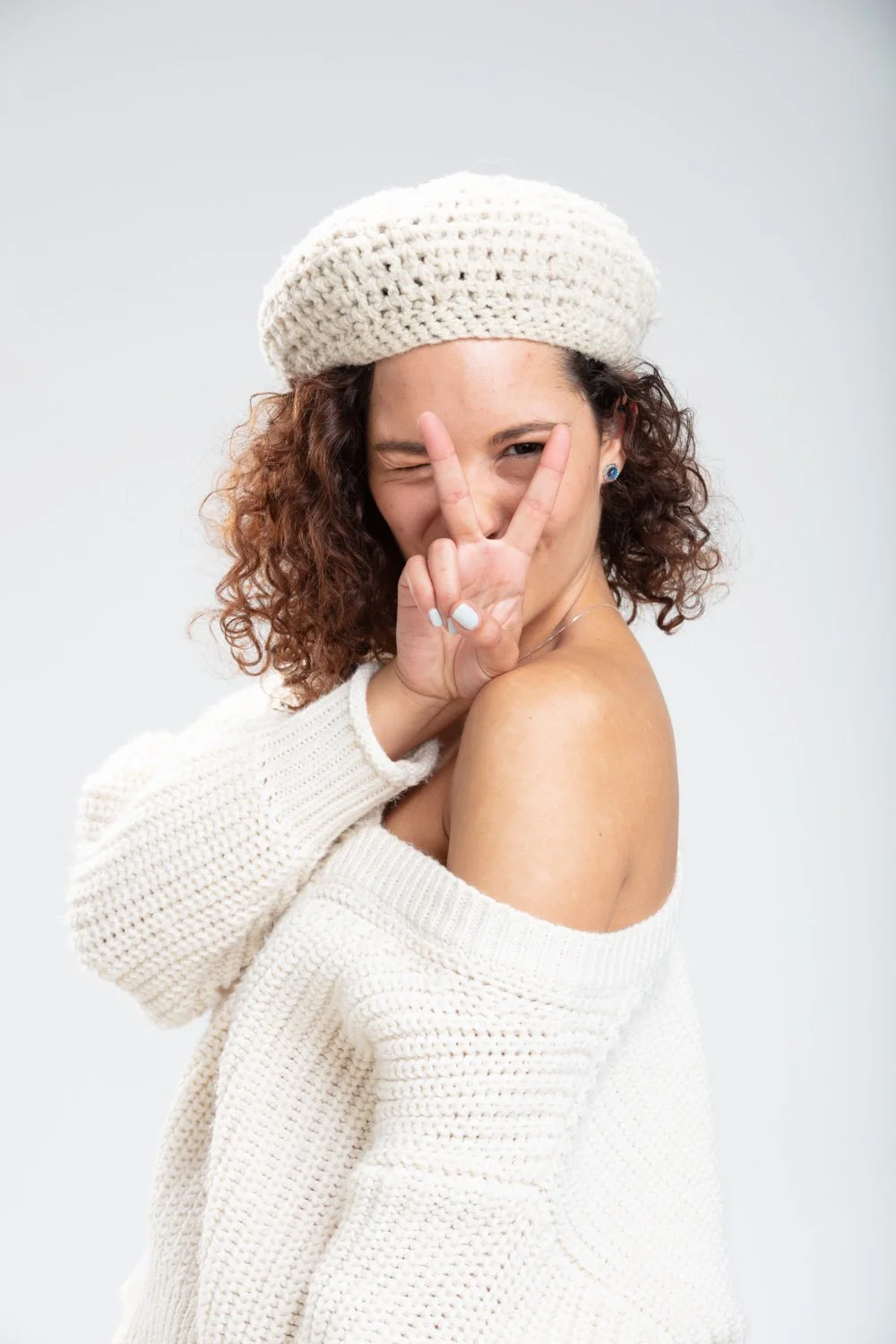 Mujer con cabello rizado, llevando un gorro de lana beige y un suéter blanco, haciendo una señal de paz con la mano y sonriendo.