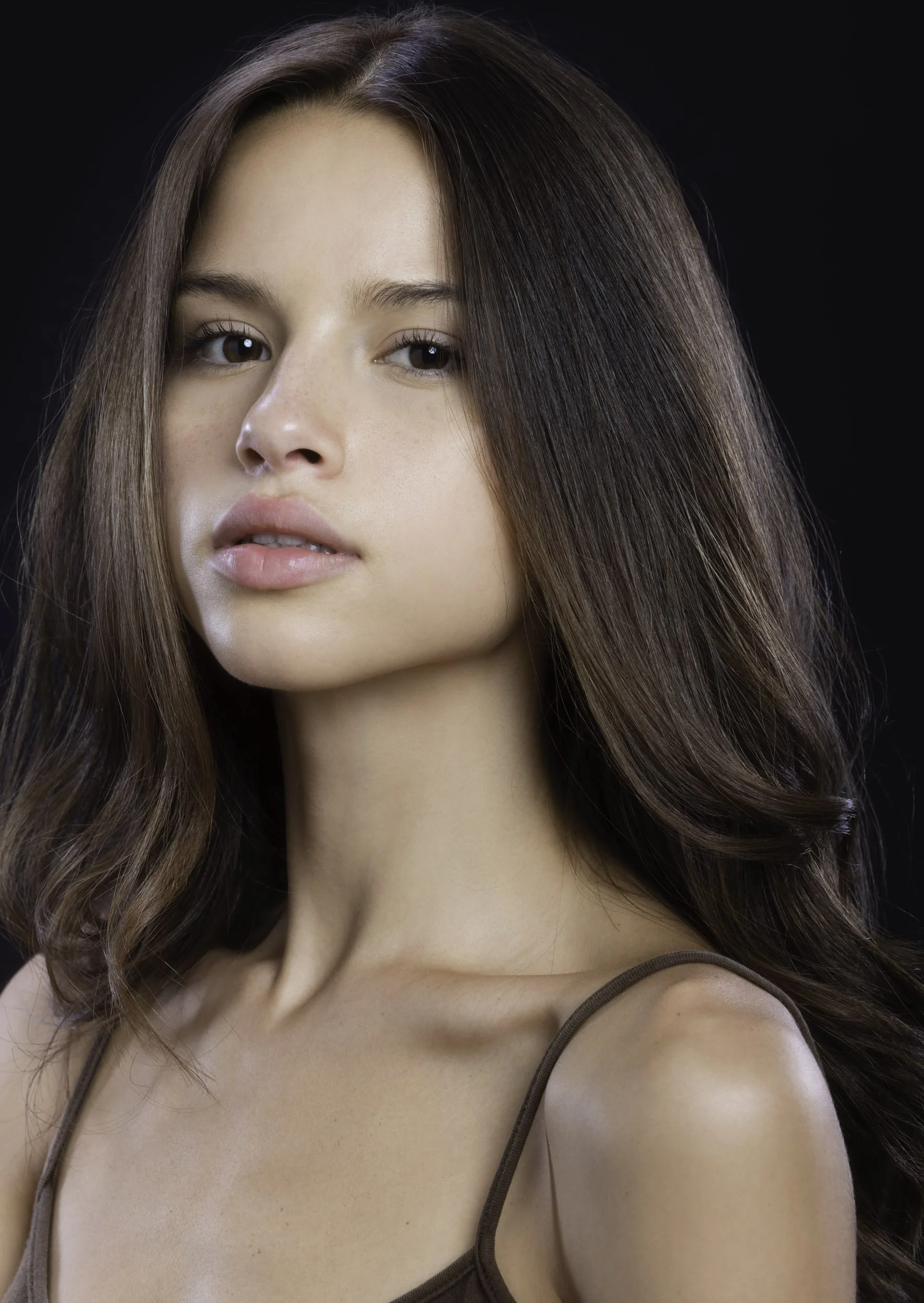 Retrato de una mujer con cabello largo y rizado, piel clara, sin maquillaje, con una expresión serena, lleva un top de tirantes delgados color marrón y fondo negro.
