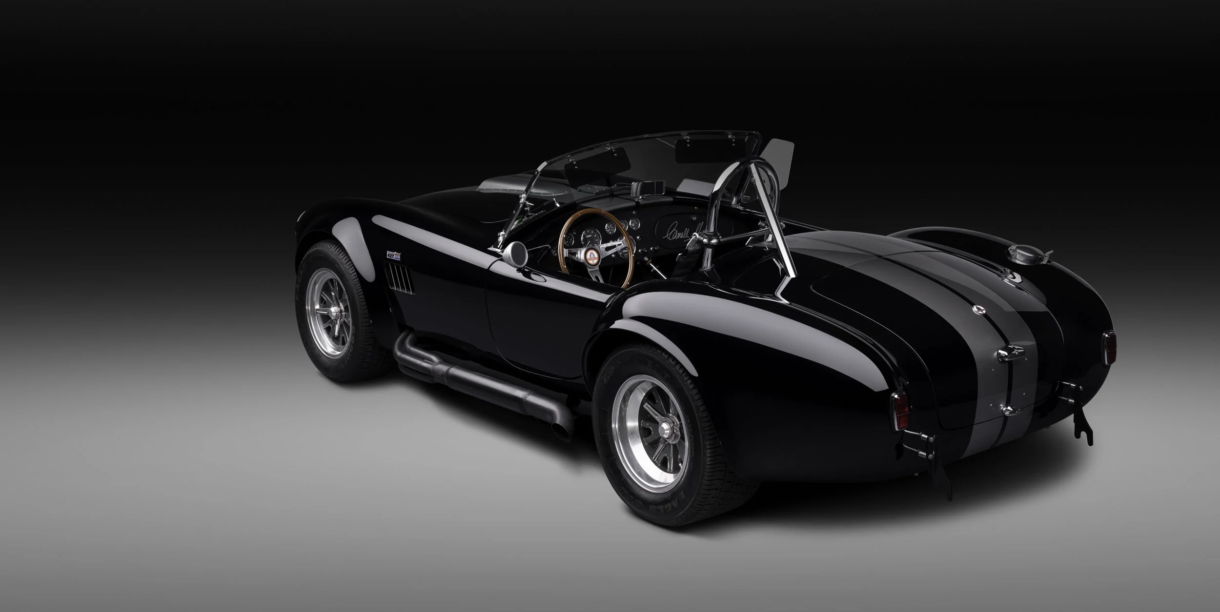 Coche deportivo clásico de color negro con detalles en gris y un interior de tablilla de madera, en un fondo negro y gris claro