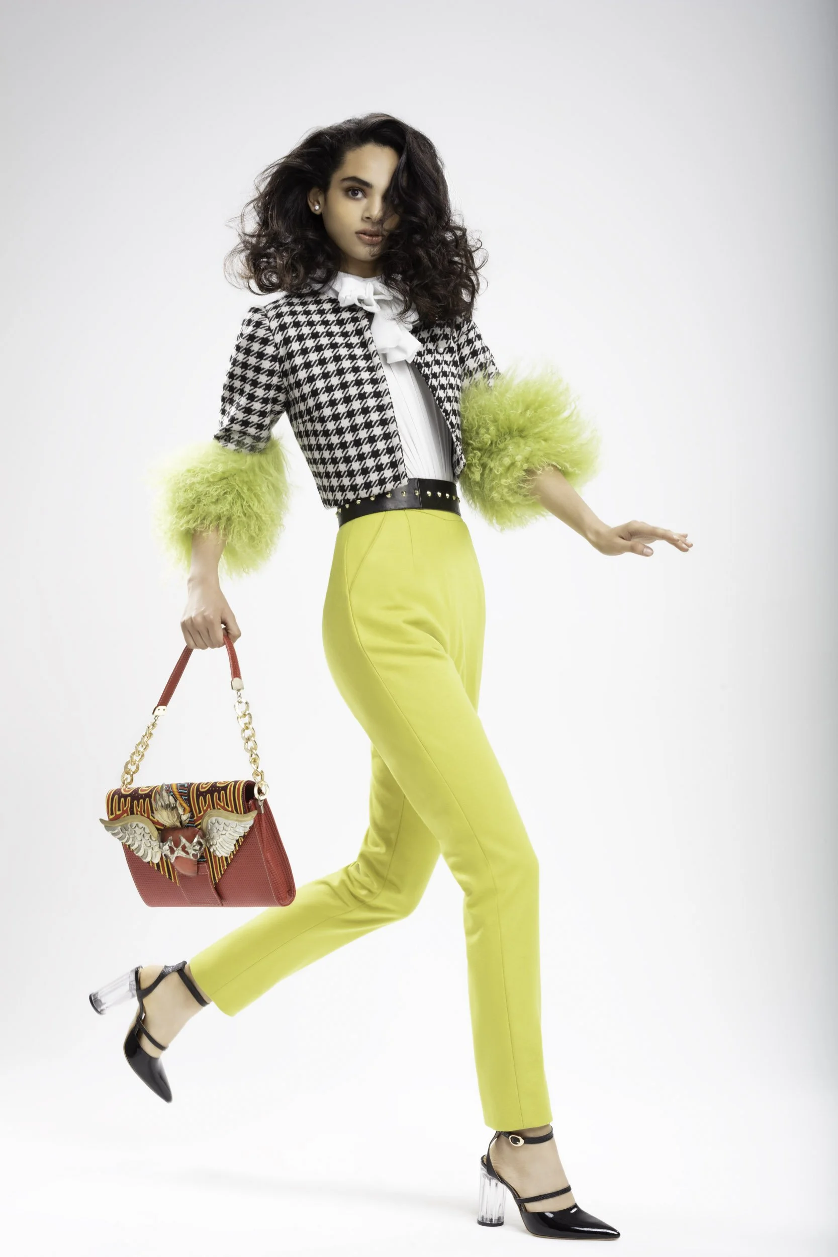 Mujer con cabello rizado y oscuro, vistiendo pantalones amarillos, blusa blanca, chaqueta a cuadros con pelos verdes en las mangas, zapatos de tacón con plataforma transparente. Sostiene un bolso color rojo con detalles y alas decorativas.