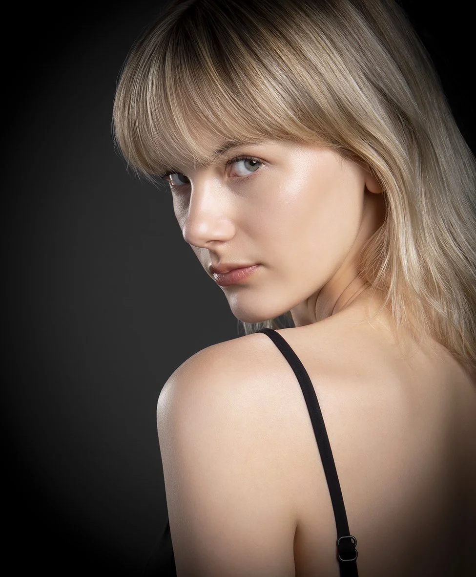 Retrato de una joven con cabello rubio y ojos claros, en un fondo oscuro, usando un vestido con una delgada correa negra.