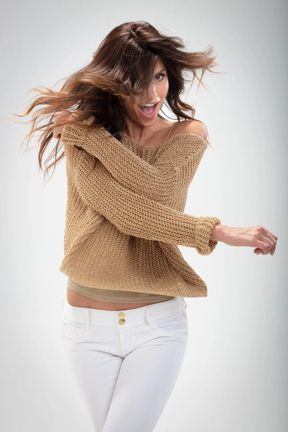Mujer sonriendo y levantando el brazo, con cabello rubio y suelto, usando un suéter color arena y pantalones blancos.