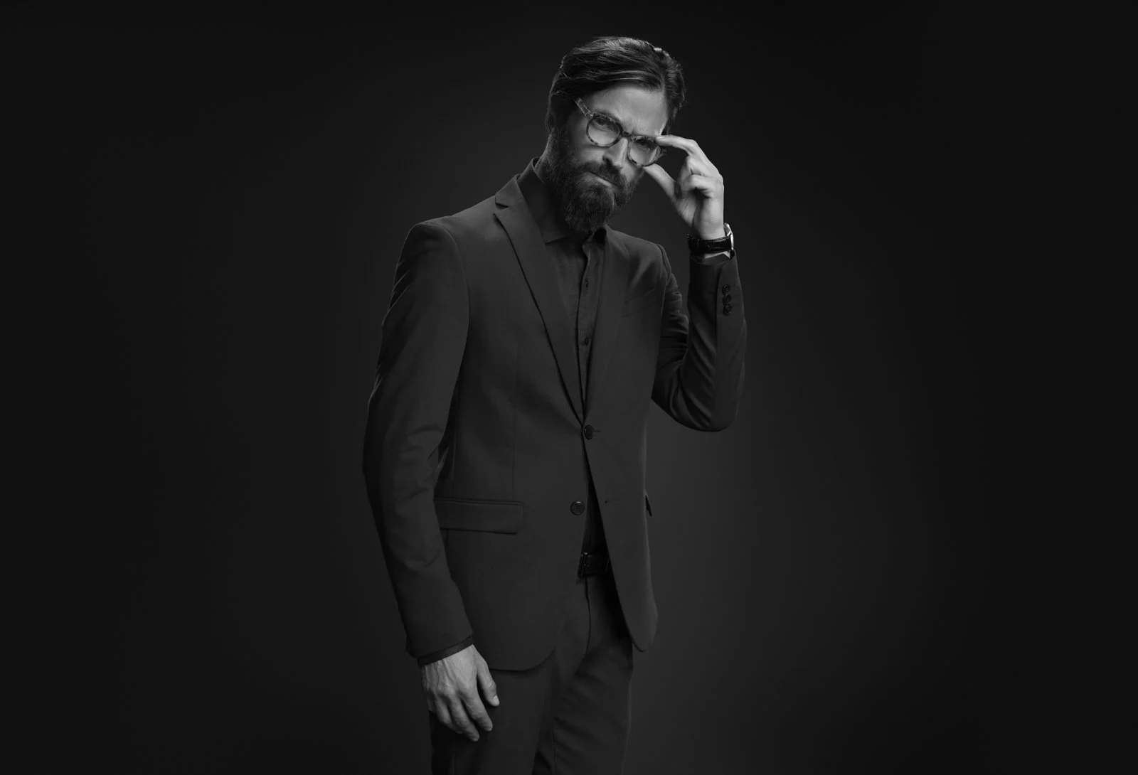 Hombre de cabello oscuro y barba, vestido con traje oscuro y gafas, posando con expresión pensativa en un fondo negro.