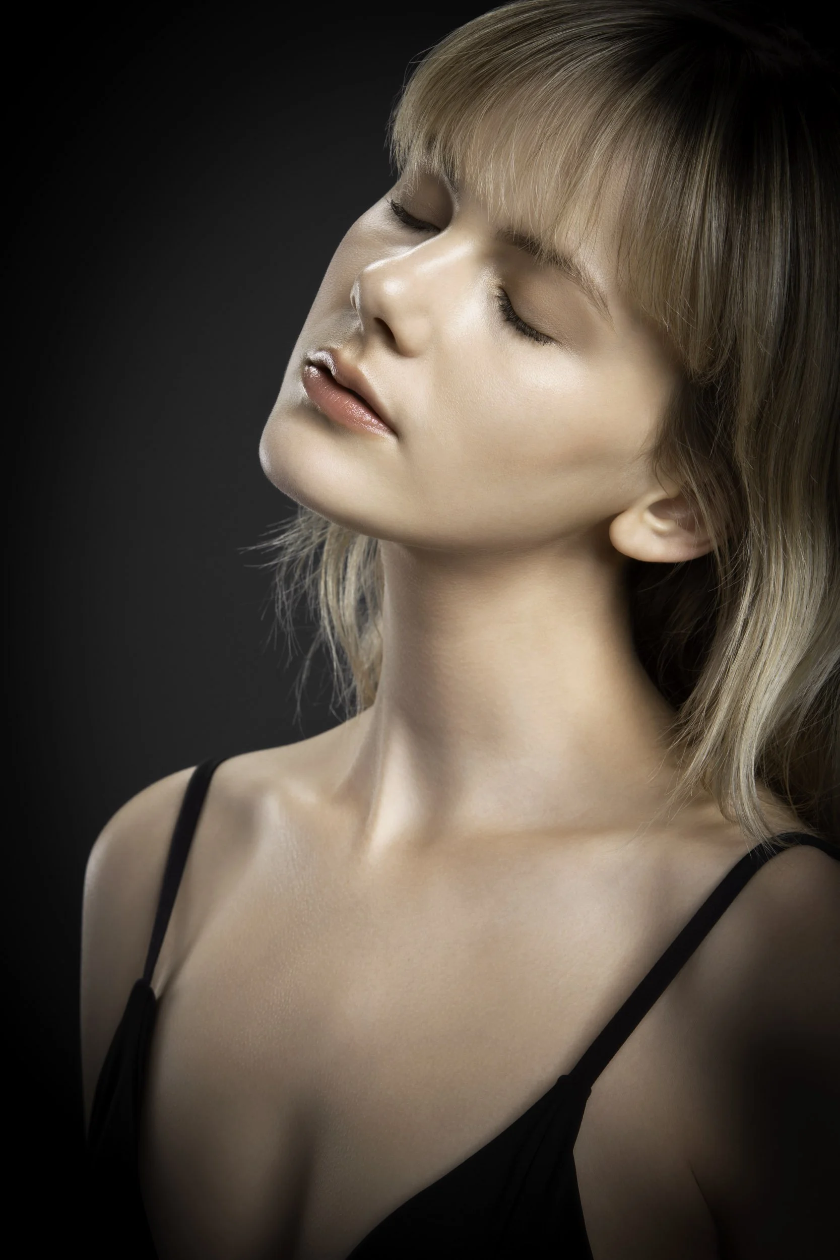 Retrato de una mujer con cabello rubio, ojos cerrados, usando un vestido negro con tirantes finos, en un fondo oscuro.