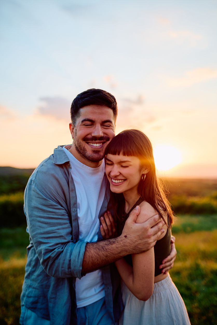 A happy couple hugging outdoors during sunset with the sun setting behind them in a field.