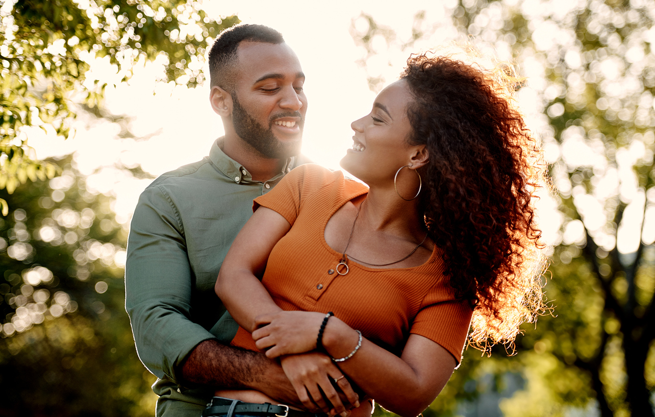 A joyful couple smiling and looking at each other outdoors during sunset, surrounded by trees.