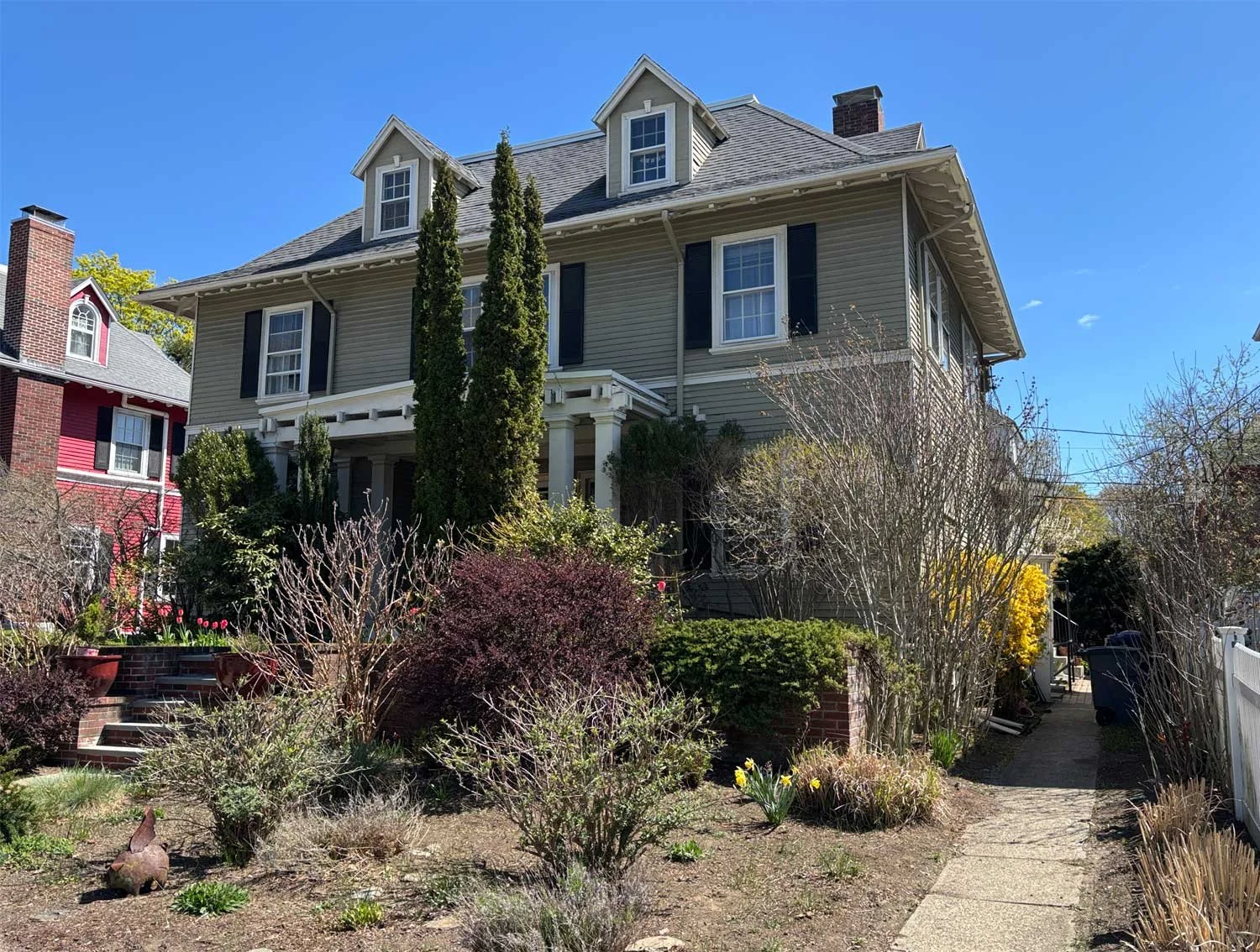 Two-story yellow house in Jamaica Plain with white trim and black shutters, featuring a front porch with red door, dormer windows on the roof, and surrounded by a yard with trees.