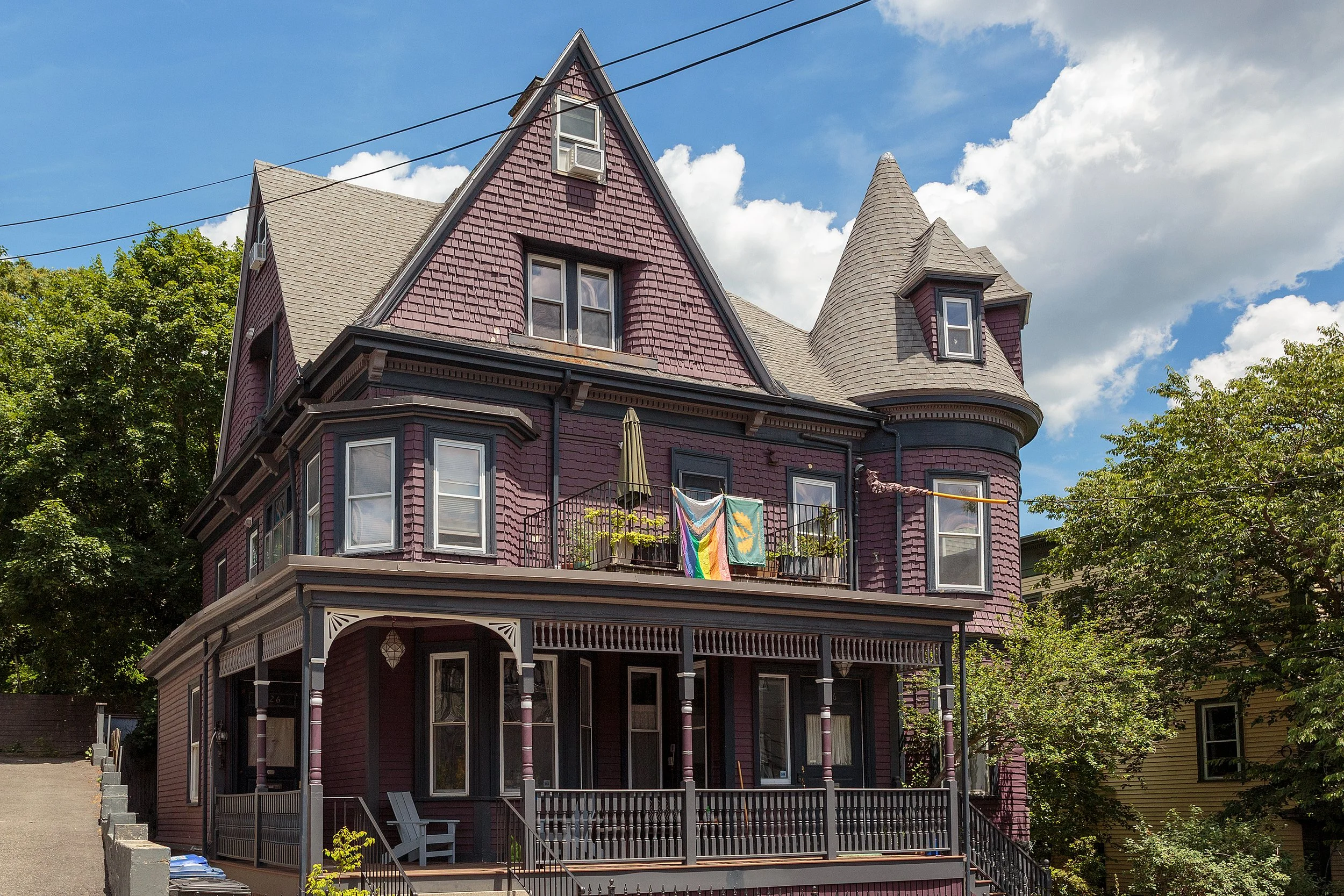 A red two-story house in Jamaica Plain with a peaked roof, surrounded by greenery and trees, features a brick pathway leading to its front entrance.