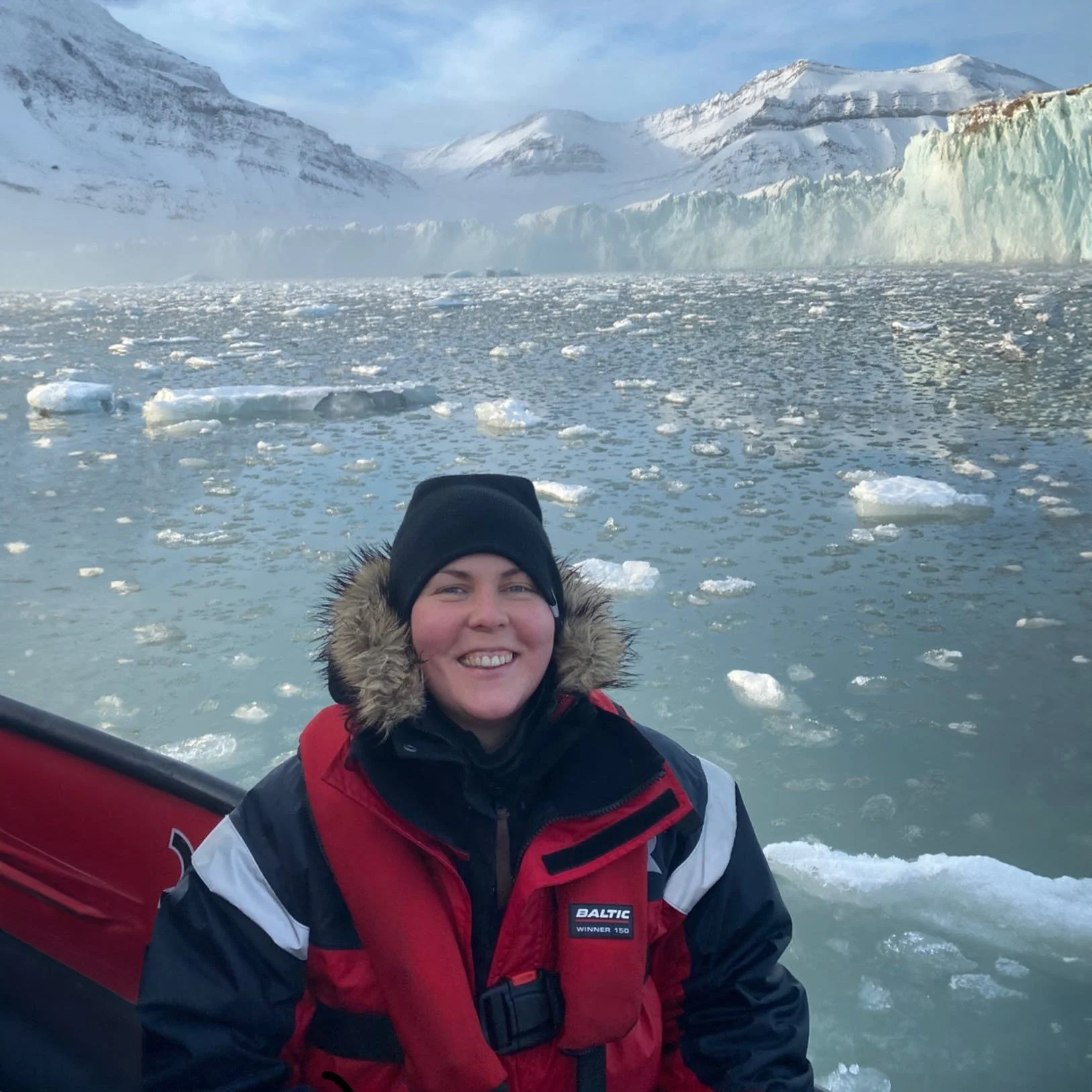 En person i kall vinterkläder på en båt, med en isberg- och snöklädd fjäll i bakgrunden, omgiven av glittrande istäckt vatten.