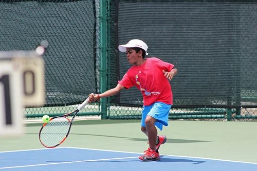 Young boy playing tennis on an outdoor court, wearing a red shirt, blue shorts, and a white cap, hitting a tennis ball.