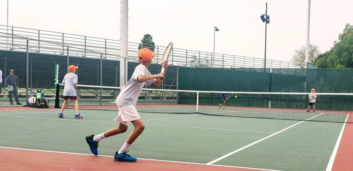 Four children playing tennis on an outdoor court, with one boy preparing to hit the ball while the others are positioned around the court.