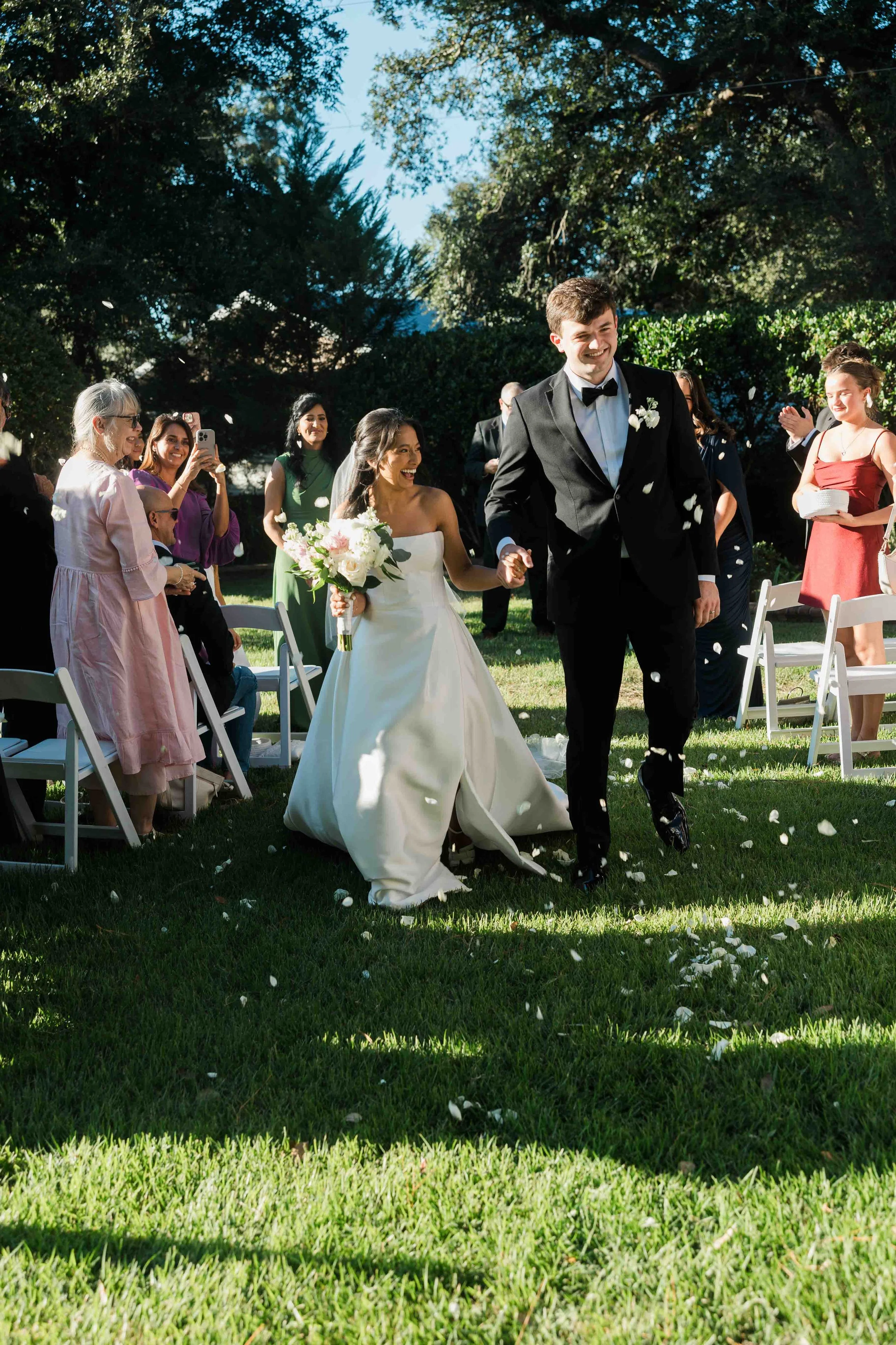 Bride and groom smile walking down the aisle with flower petals thrown at them, they are smiling.