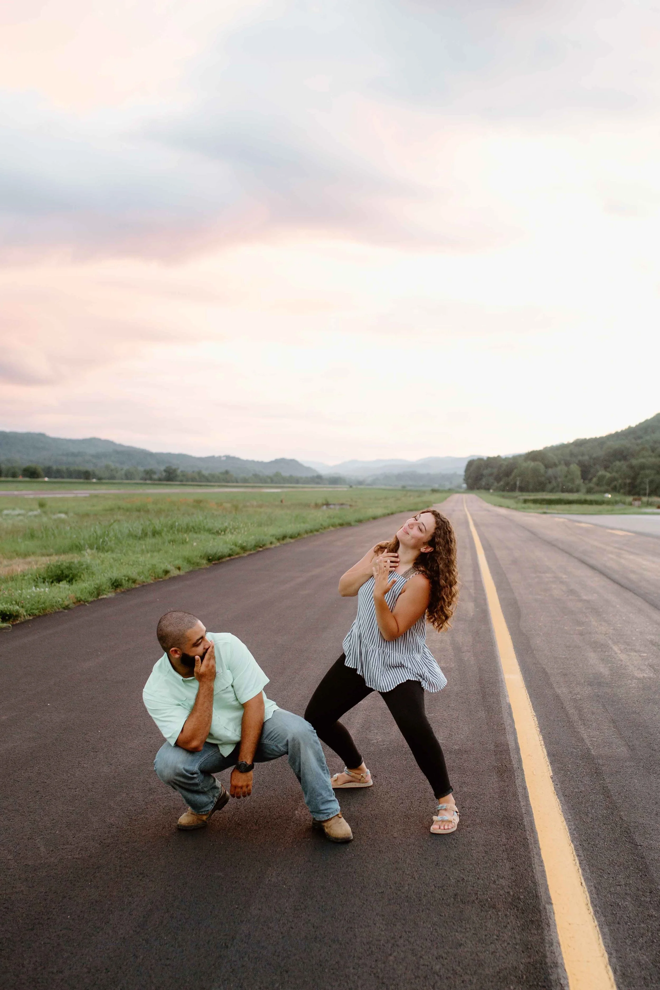 Two people posing playfully on a road with an expansive landscape and colorful sky in the background.