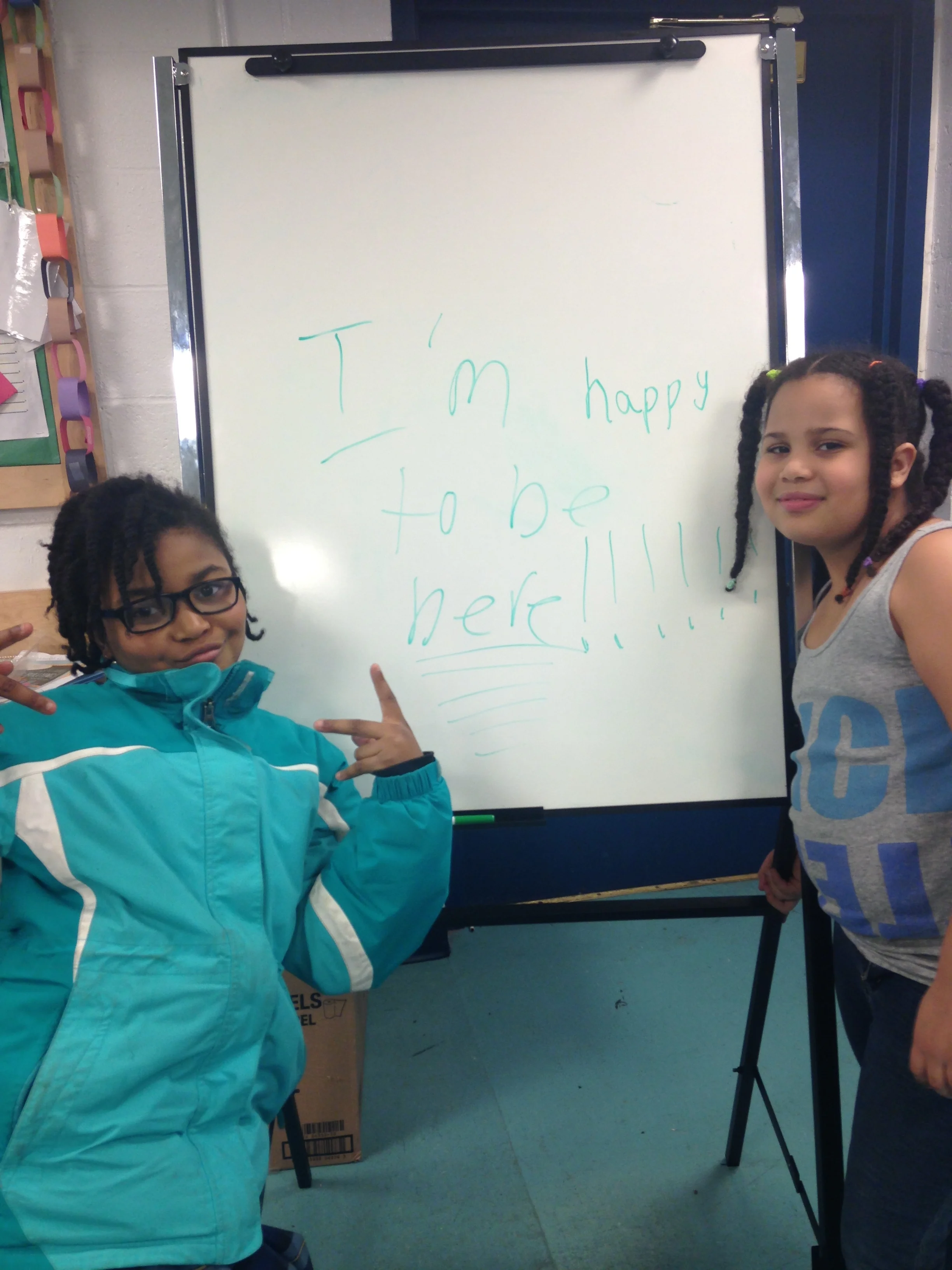 Two students pose with their writing on a whiteboard: "I'm happy to be here!!!!!!"