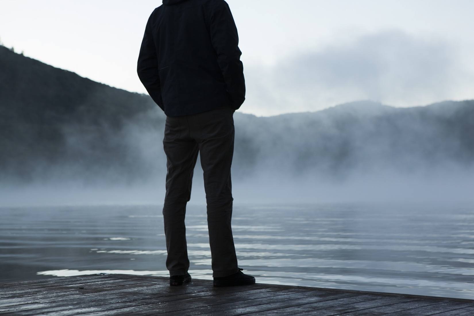 Man standing on dock overlooking a lake and mountains in the mist contemplating life