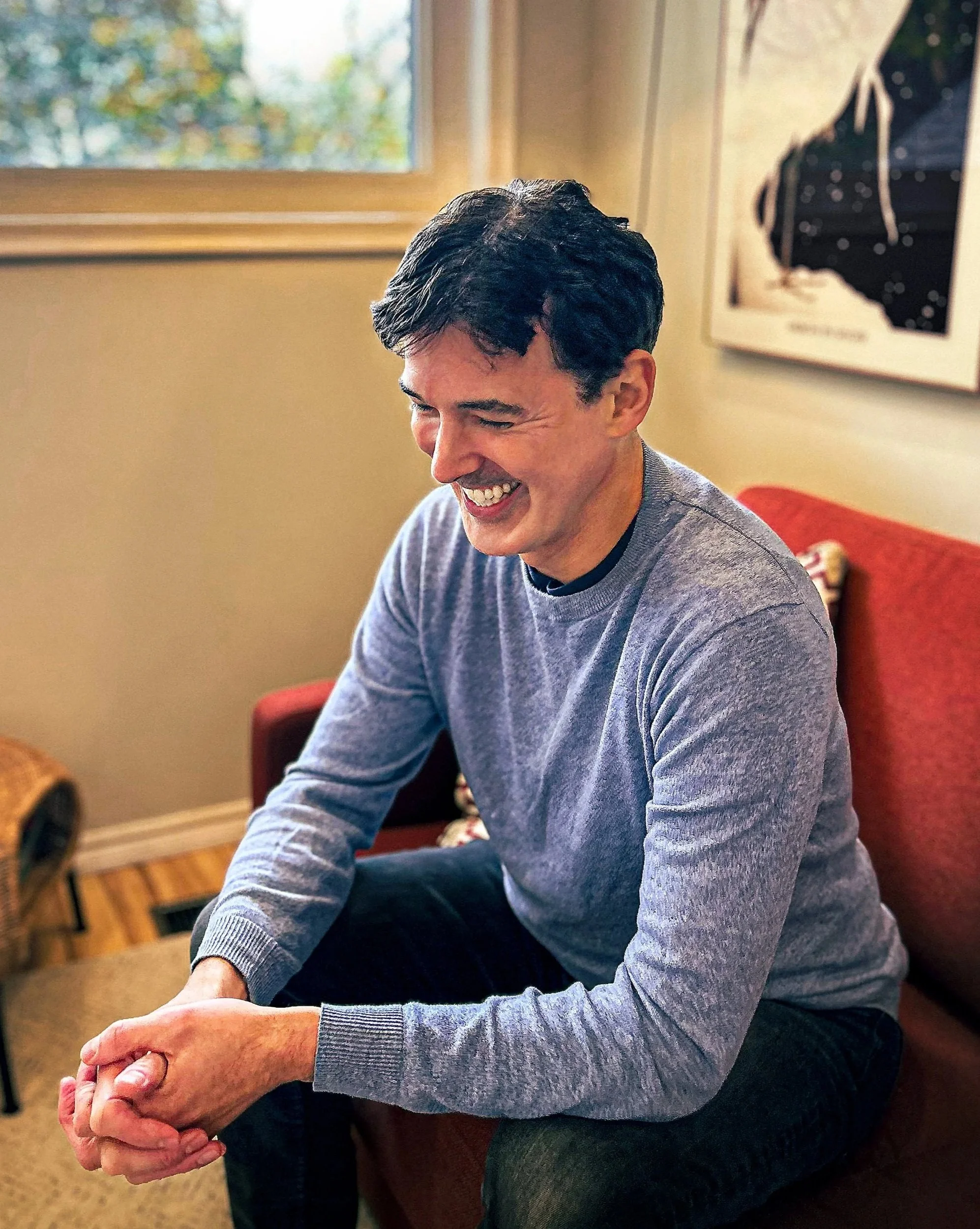 Portrait of therapist Nat Roman, sitting on a red couch, looking down and smiling.