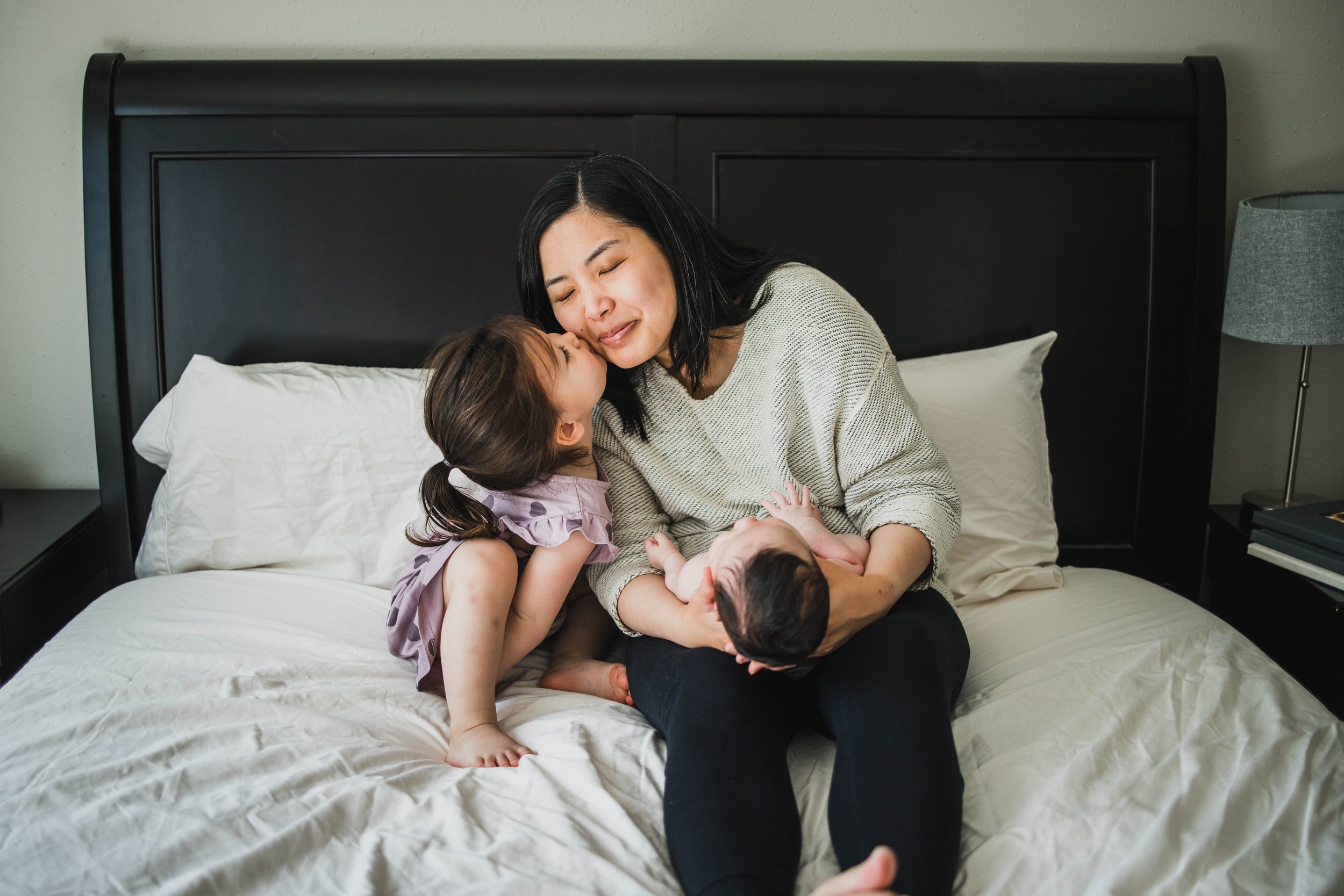 Toddler daughter leans up to kiss her mother on the cheek as she cradles the naked newborn. An emotional moment from an in-home seattle newborn photography session.