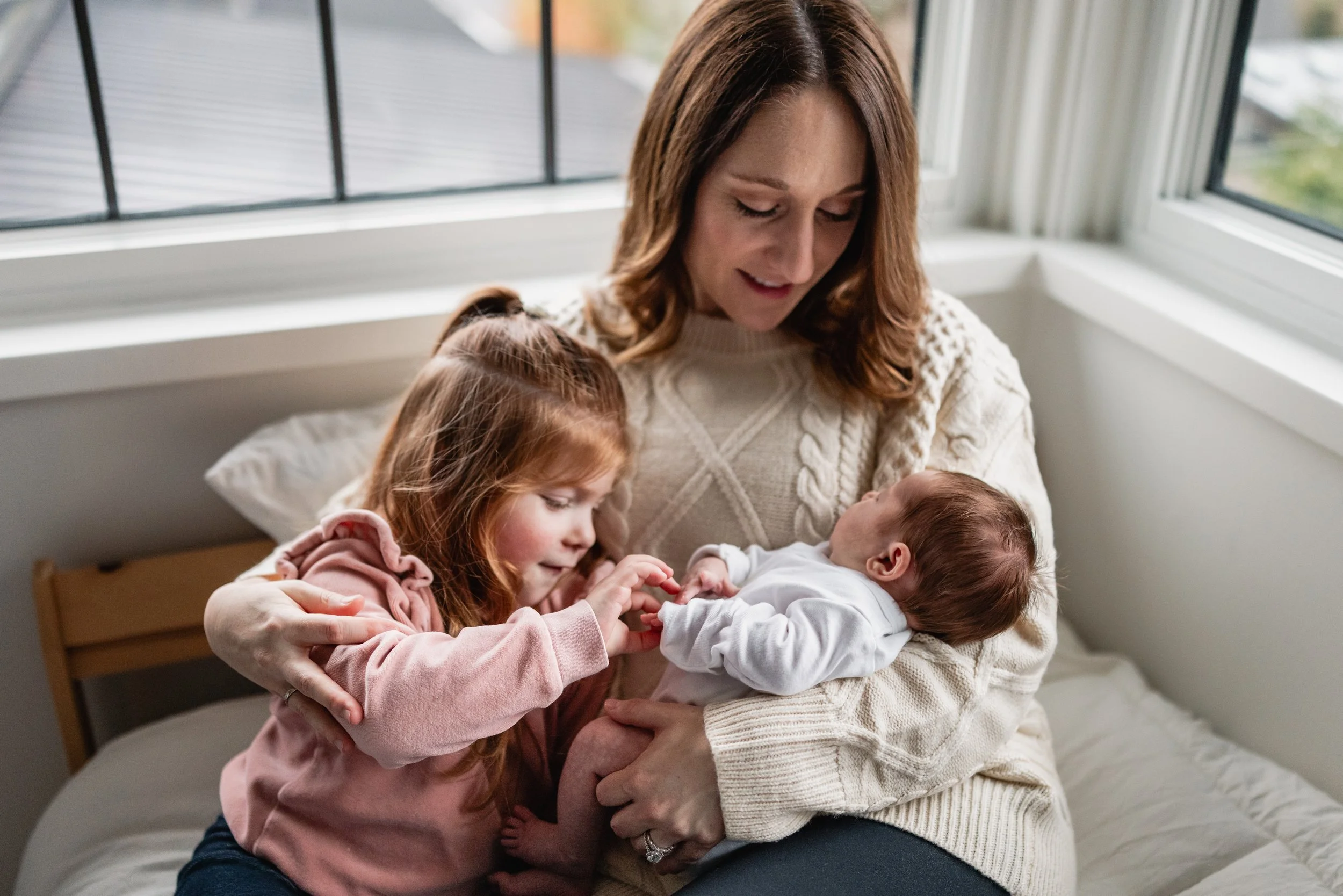 Mom snuggles with her toddler and newborn in the corner window of her Seattle home.