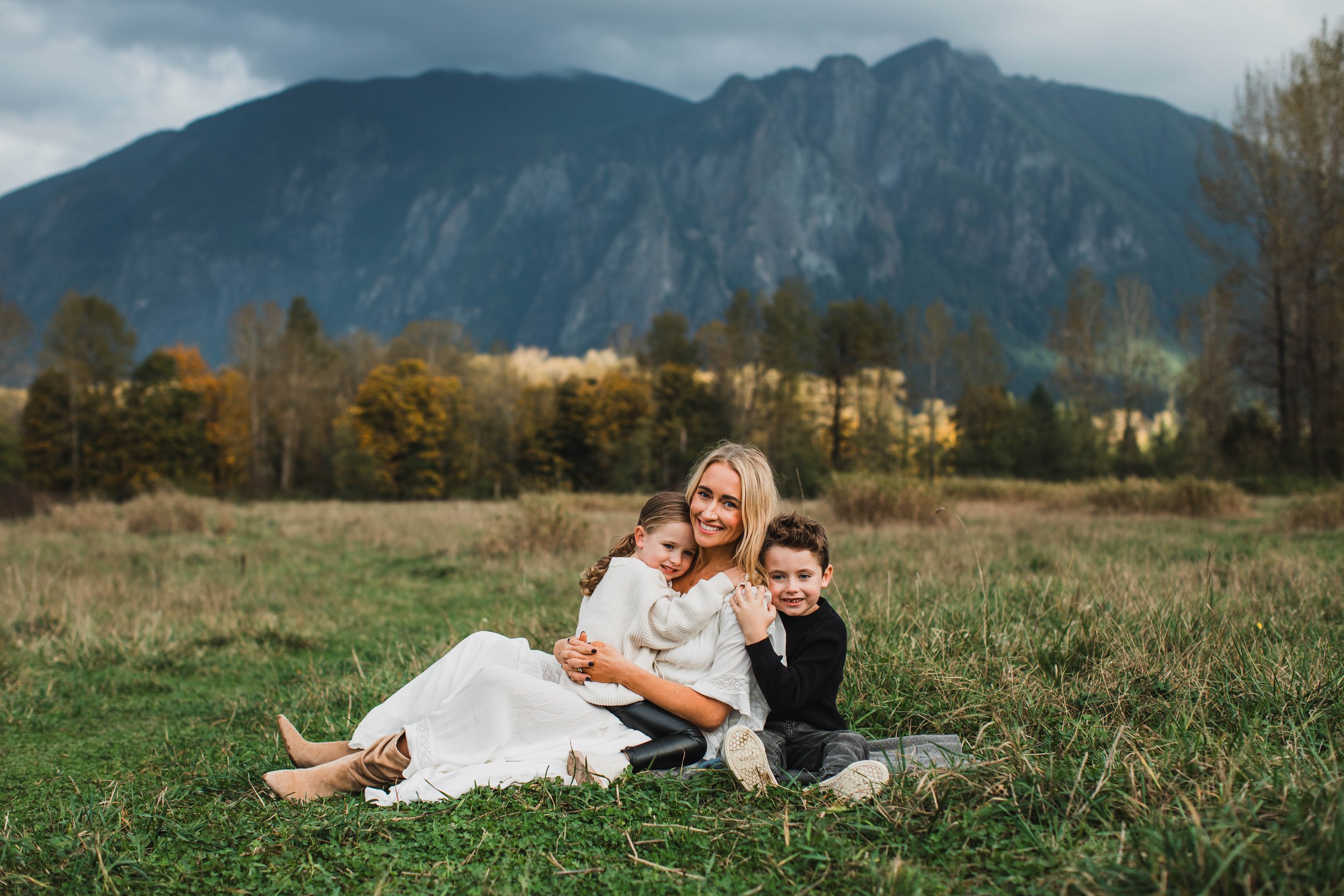 Seattle family photography in a large grassy field with a view of Mount Si. Mother sits with children for an outdoor portrait session in North Bend.