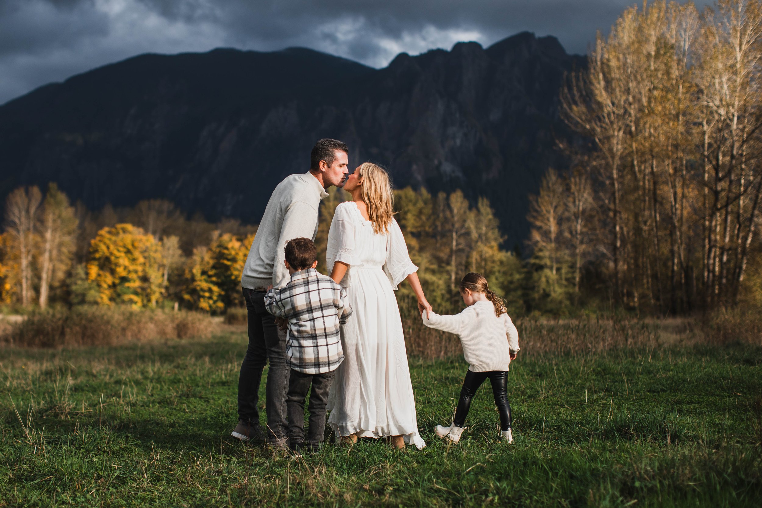 Parents kiss as their children play in a grassy meadow in front of Mount Si, featured in a North Bend Seattle family photography shoot.
