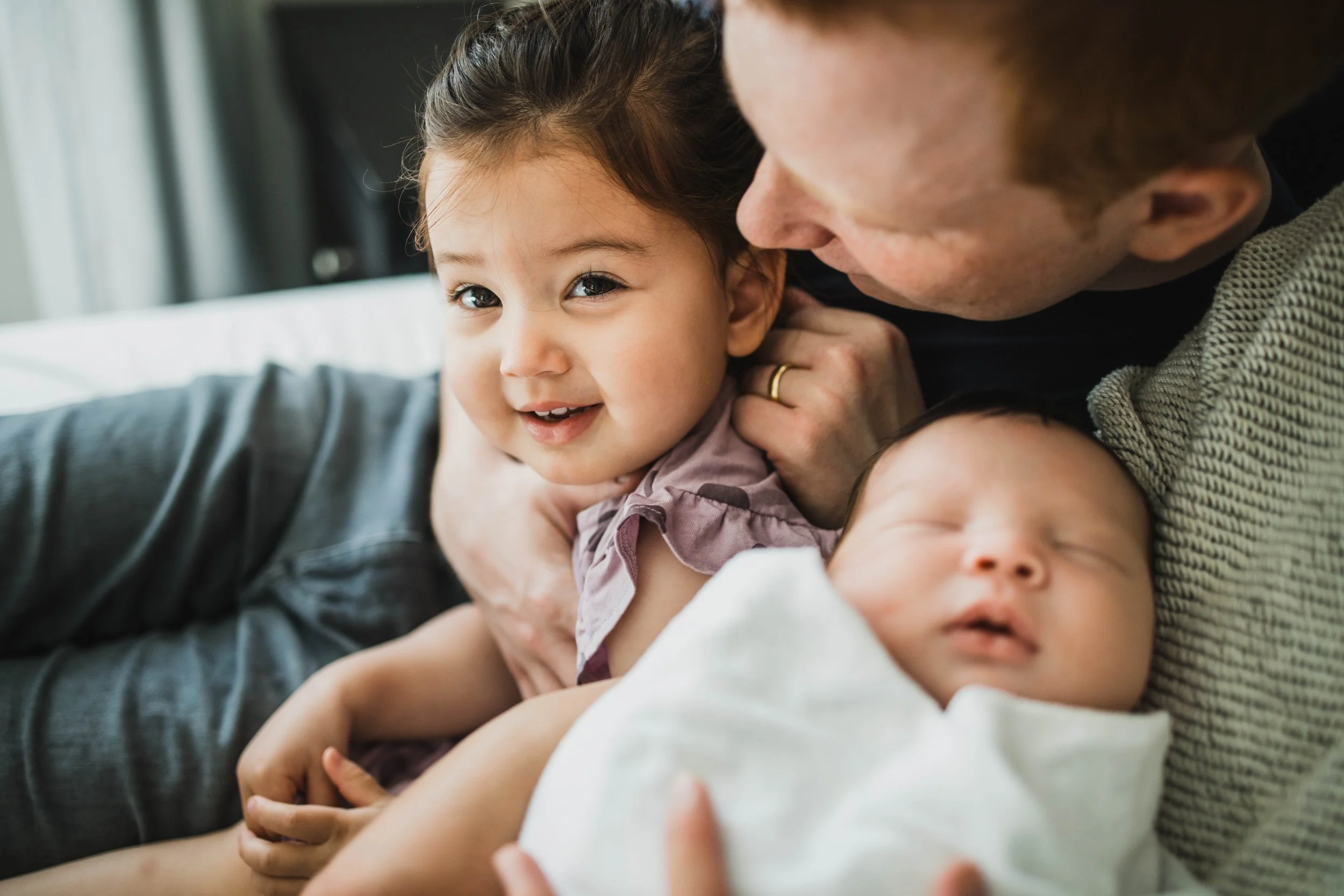 A toddler girl looks at the camera while her father leans over to look at her sleeping baby brother. A professional in-home seattle newborn photography shot.