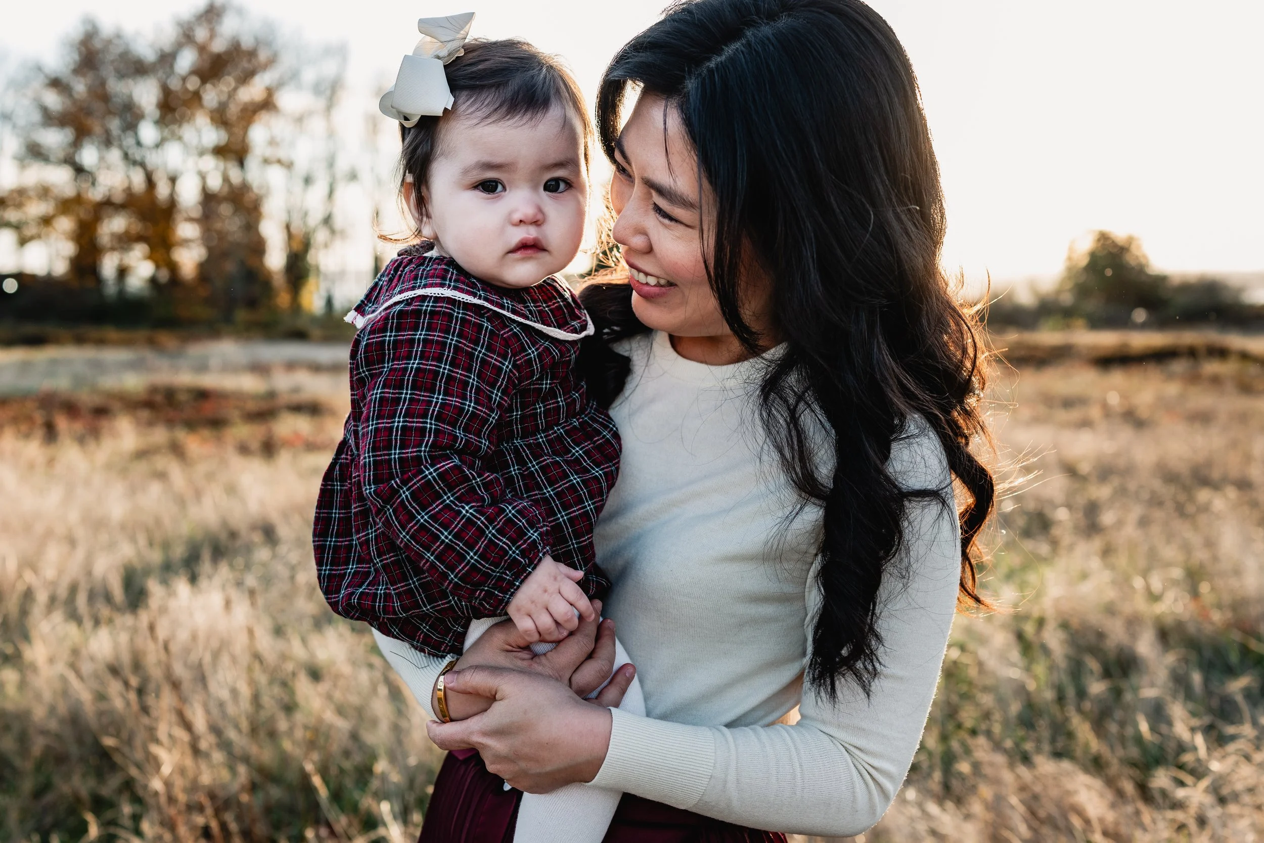 A mother smiles warmly at her baby daughter during an autumnal Seattle family photography session. The baby wears a coordinating fall plaid dress.