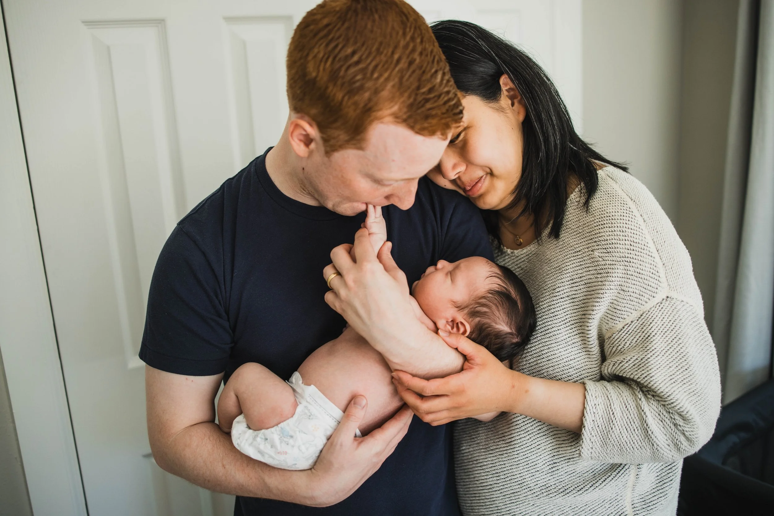 Parents share an intimate moment looking down at their sleeping newborn with dark hair, standing in their nursery. A candid in-home seattle newborn photography portrait.