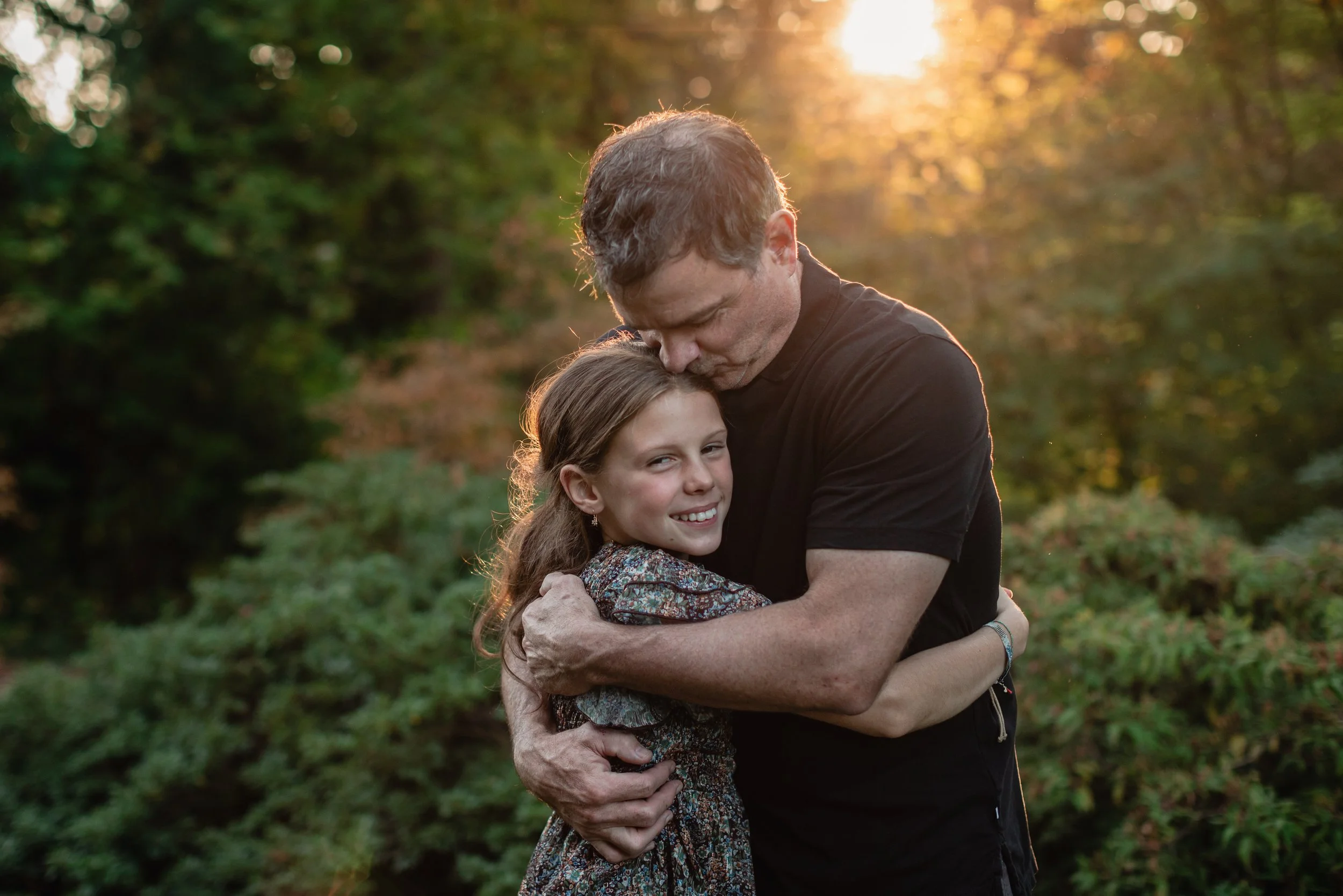 A father kisses his smiling daughter’s head as they hug in a sun-drenched garden at the Seattle Arboretum during a Seattle family photography session.