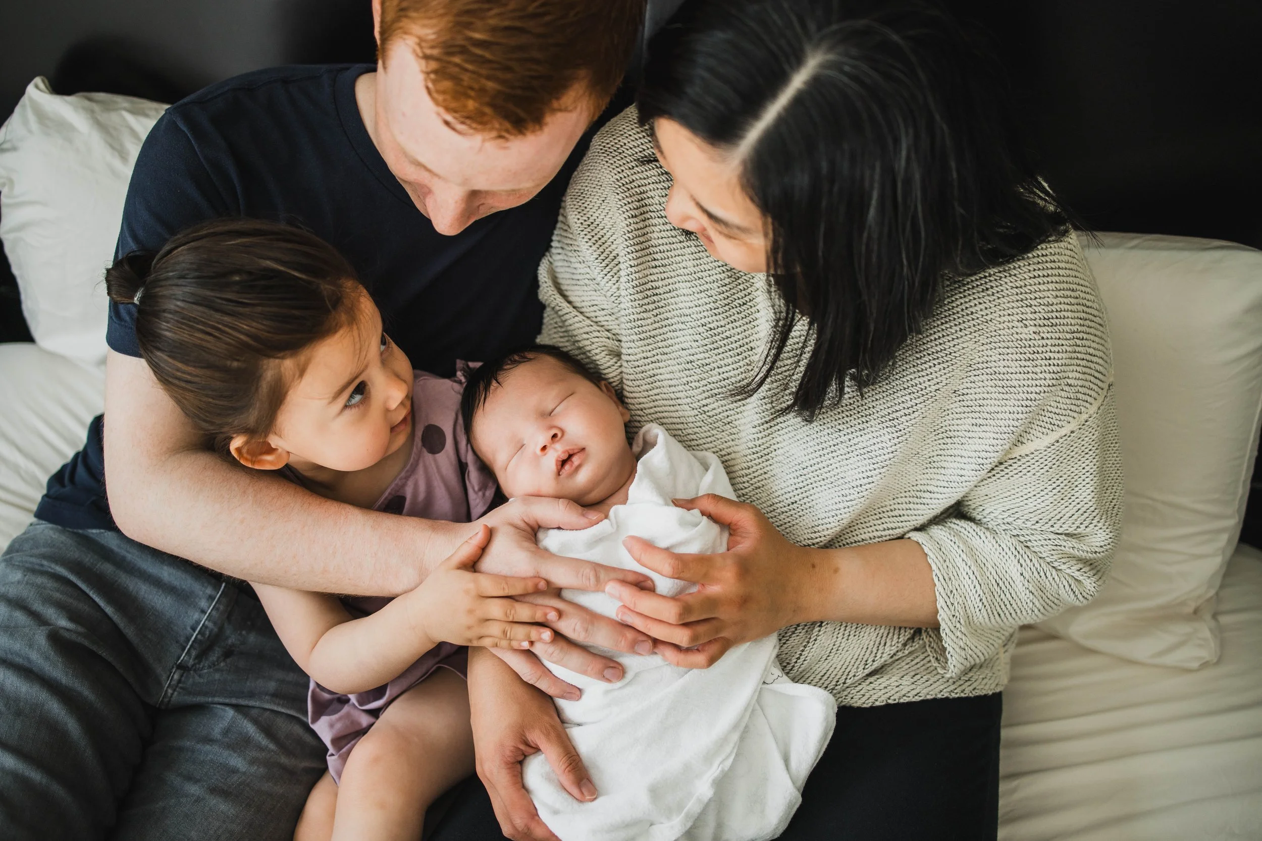 An overhead view of parents and their toddler daughter gazing down at the sleeping newborn in a collective embrace. In-home seattle newborn photography.