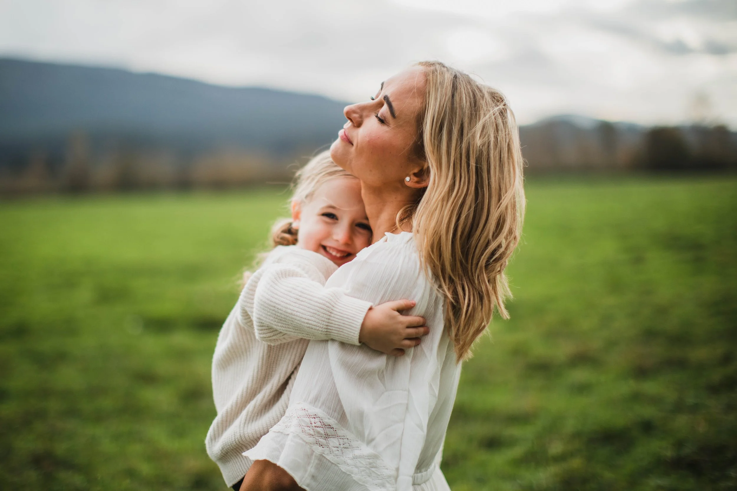 An intimate moment between a mother and daughter, hugging in a green field during a North Bend Seattle family photography session. The daughter smiles at the camera while the mother smiles upwards with her eyes closed.