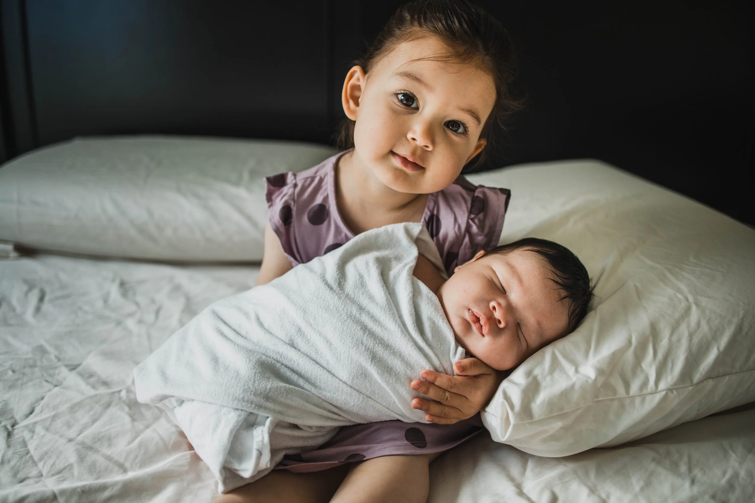A toddler girl in a polka dot dress holds her sleeping baby brother on a bed during an in-home seattle newborn photography session.